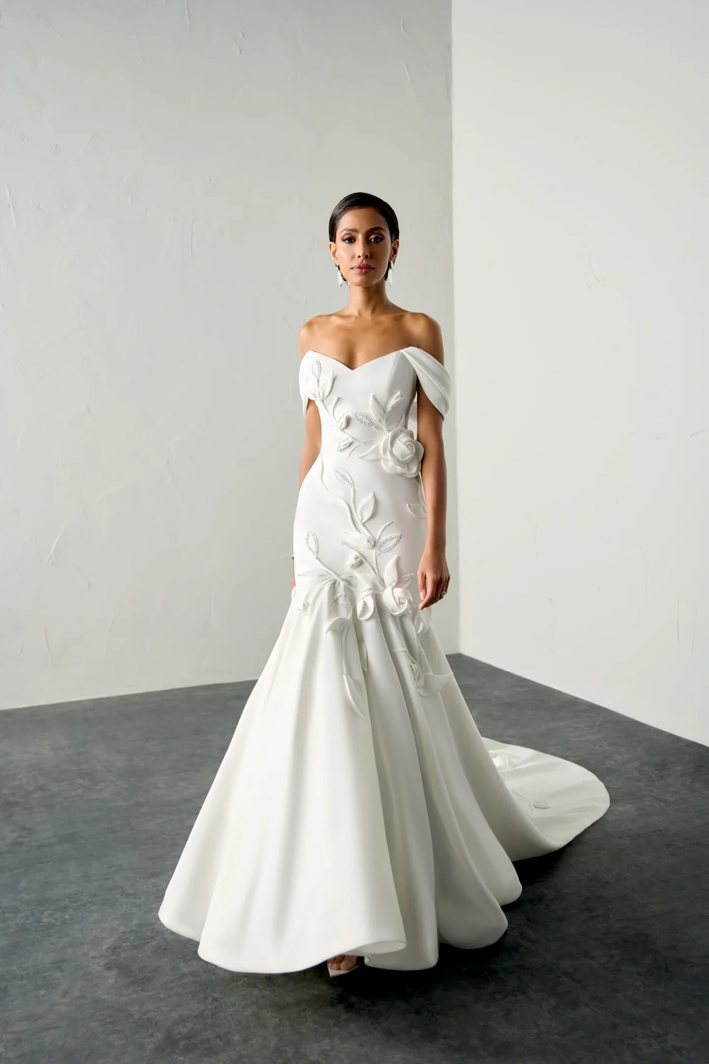 A woman in a white wedding gown with floral embroidery and off-the-shoulder sleeves, standing in a plain room with white walls and gray carpet.