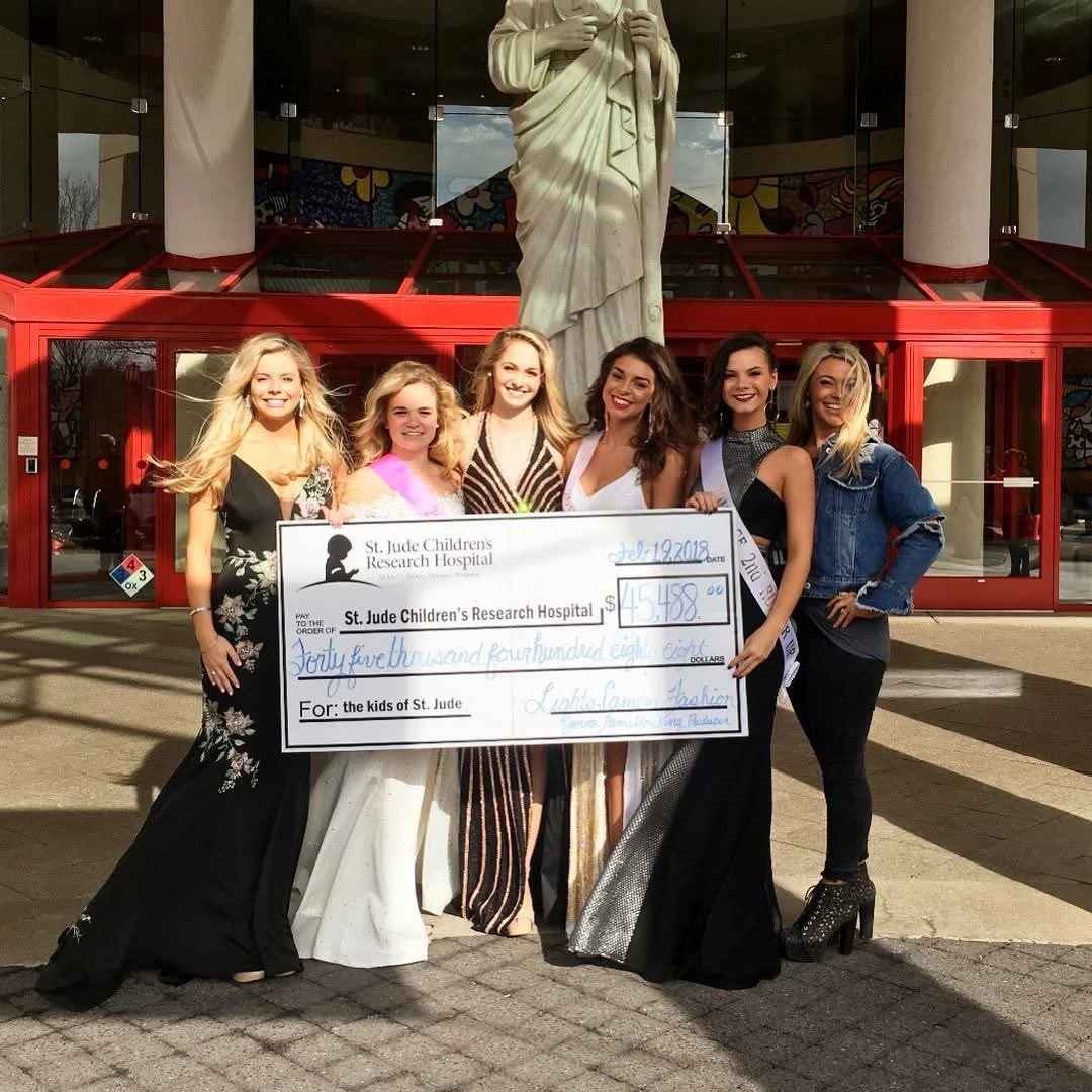 A group of seven women standing outside a building holding a large check for $45,458 donation to St. Jude Children's Research Hospital. They are dressed in formal and fashionable attire and are smiling at the camera.