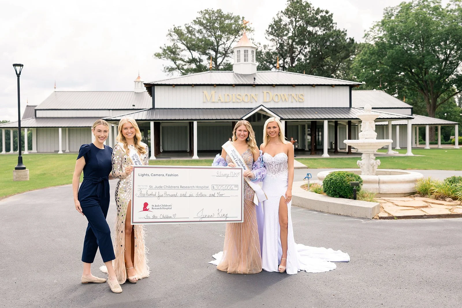 Four women standing in front of a building named 'Madison Downs,' holding an oversized check for $305,000 made out to St. Jude Children's Research Hospital. The women are dressed in formal attire, with two wearing evening gowns and one wearing a jump