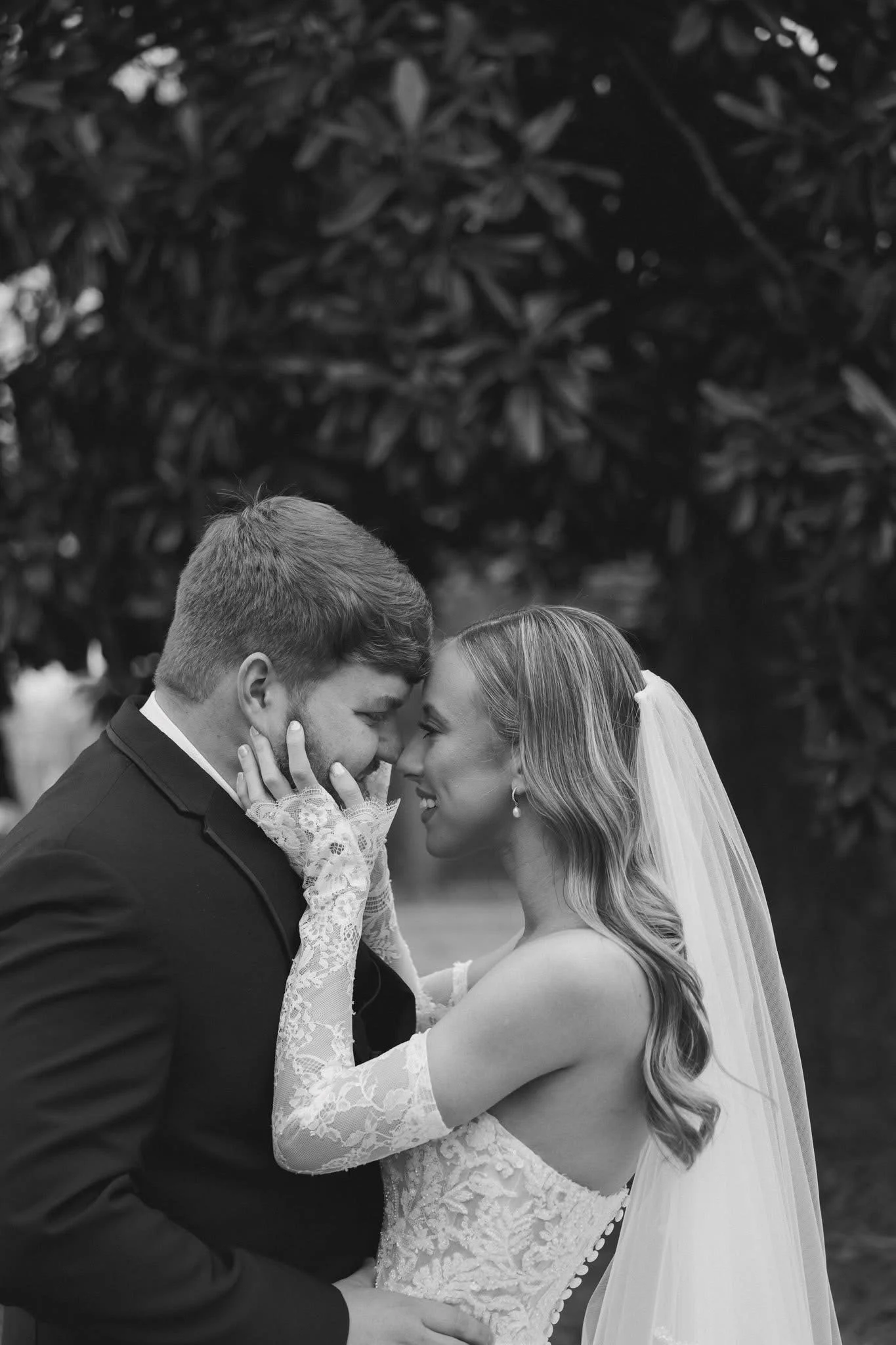 A black and white photo of a bride and groom kissing outdoors, with the groom in a tuxedo and the bride in a lace wedding dress with a veil, holding each other's faces.