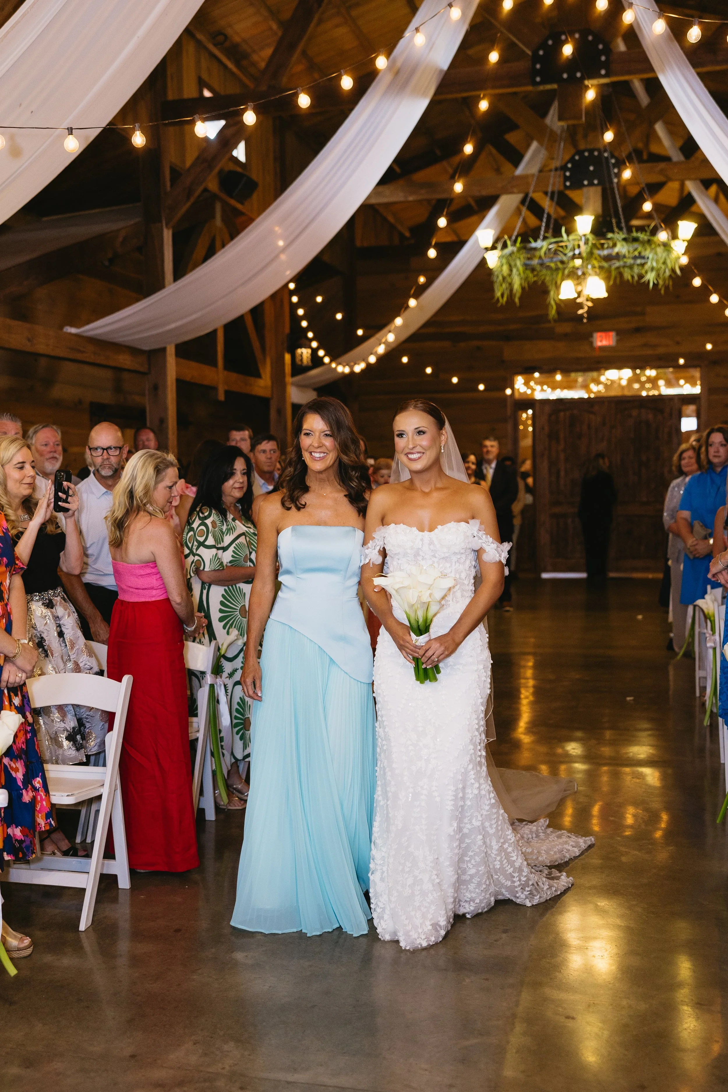 Two brides, one in a light blue strapless dress and the other in a white lace wedding gown with off-shoulder sleeves, are standing together at their wedding ceremony inside a rustic barn decorated with string lights and draped white fabric. Guests ar