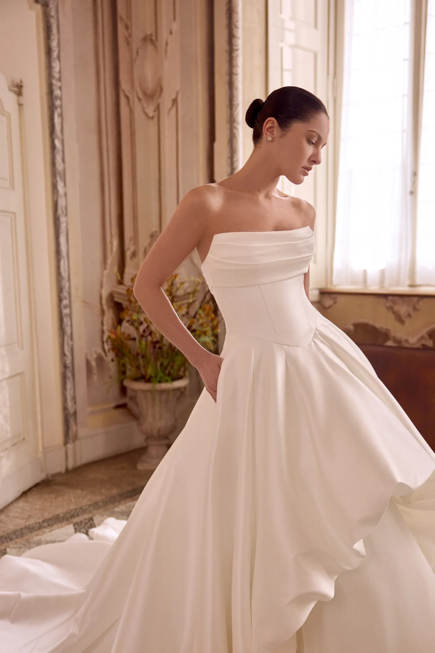 A woman in a strapless white wedding dress standing in an ornate room with detailed wall moldings and large windows, looking down.