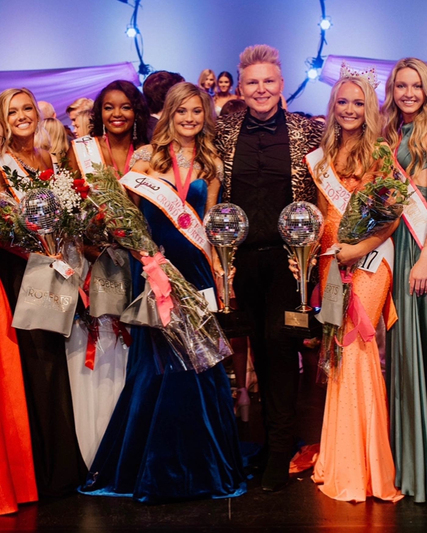 Group of women in formal dresses and sashes holding floral bouquets and awards, with a man dressed in a black shirt and leopard print blazer in the center, at a celebratory event with decorative lighting.