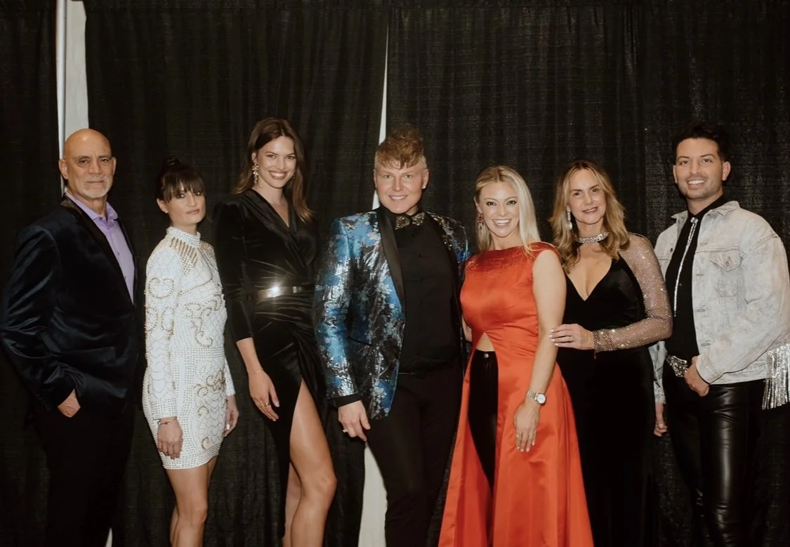 Group of eight people dressed in formal attire posing for a photo in front of black curtains.