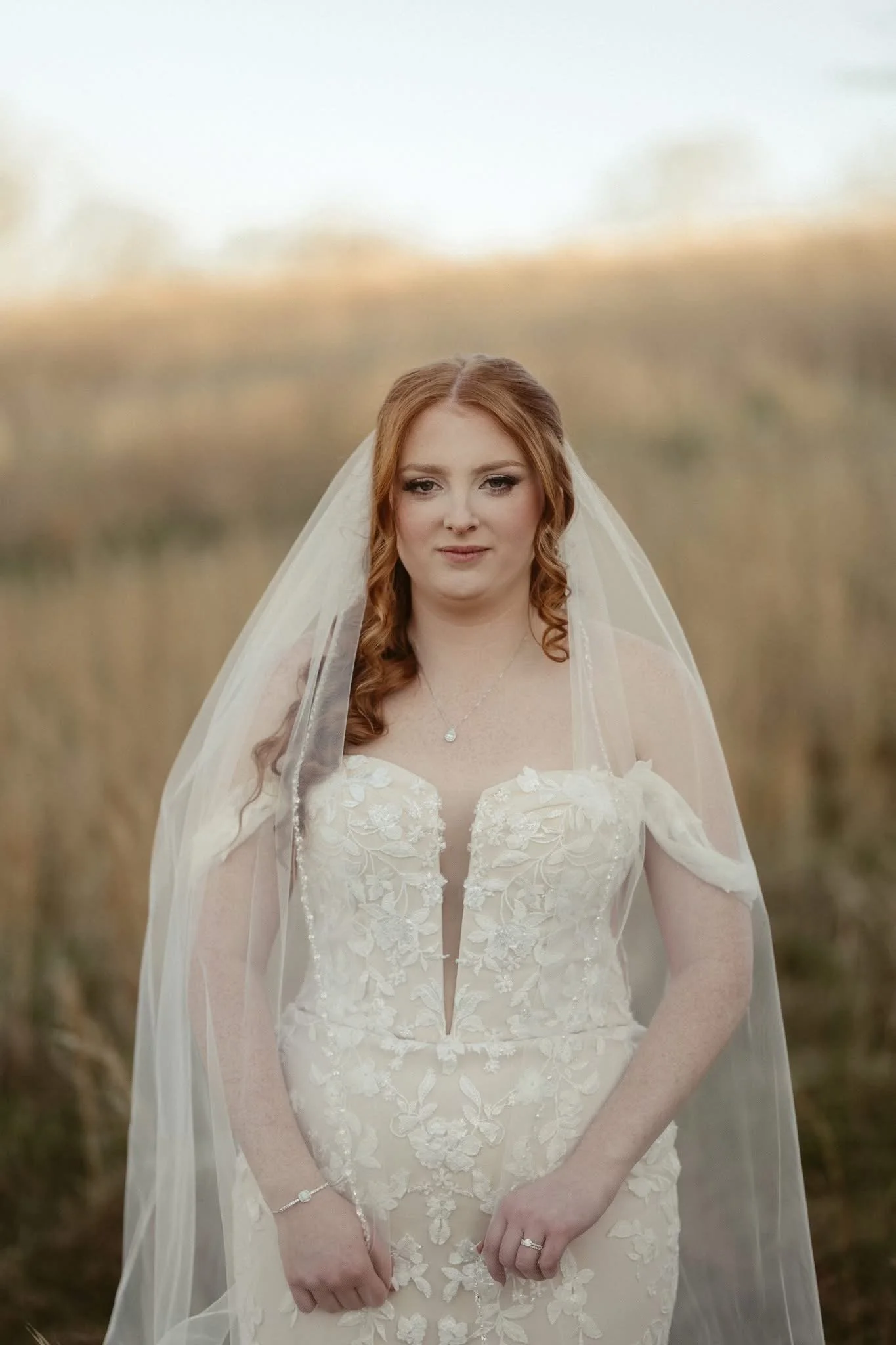 A bride with red hair wearing a white lace wedding dress and a veil standing in a field with a blurred background of grass and sky.