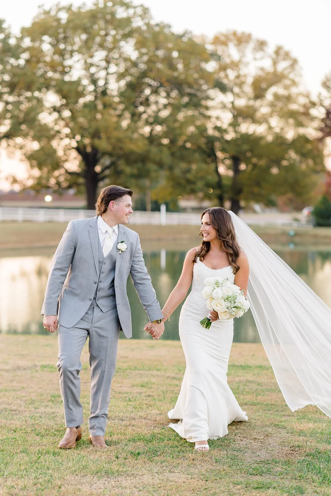 Bride and groom holding hands and walking by a lake during sunset, dressed in wedding attire, smiling at each other.