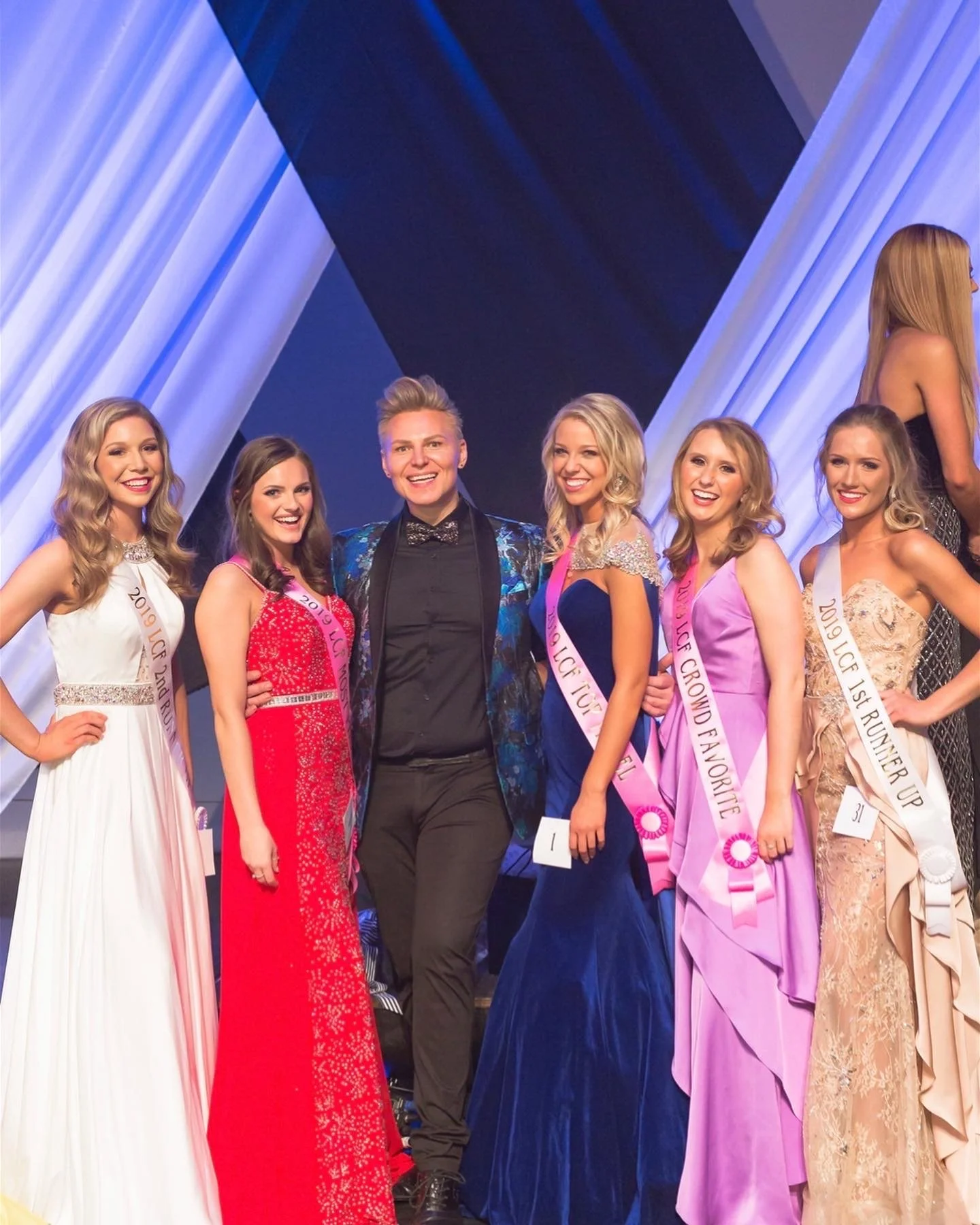 A group of women in formal dresses and sashes standing on stage at a beauty pageant, smiling. One man in the middle is dressed in a black suit with a blue patterned blazer, smiling. The women are wearing sashes with titles, and some have pink and whi