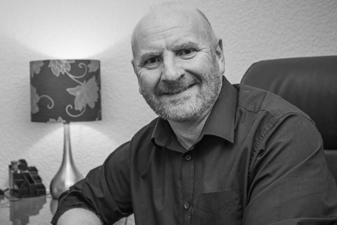 Black and white photo of a smiling man sitting at a desk with a lamp in the background.