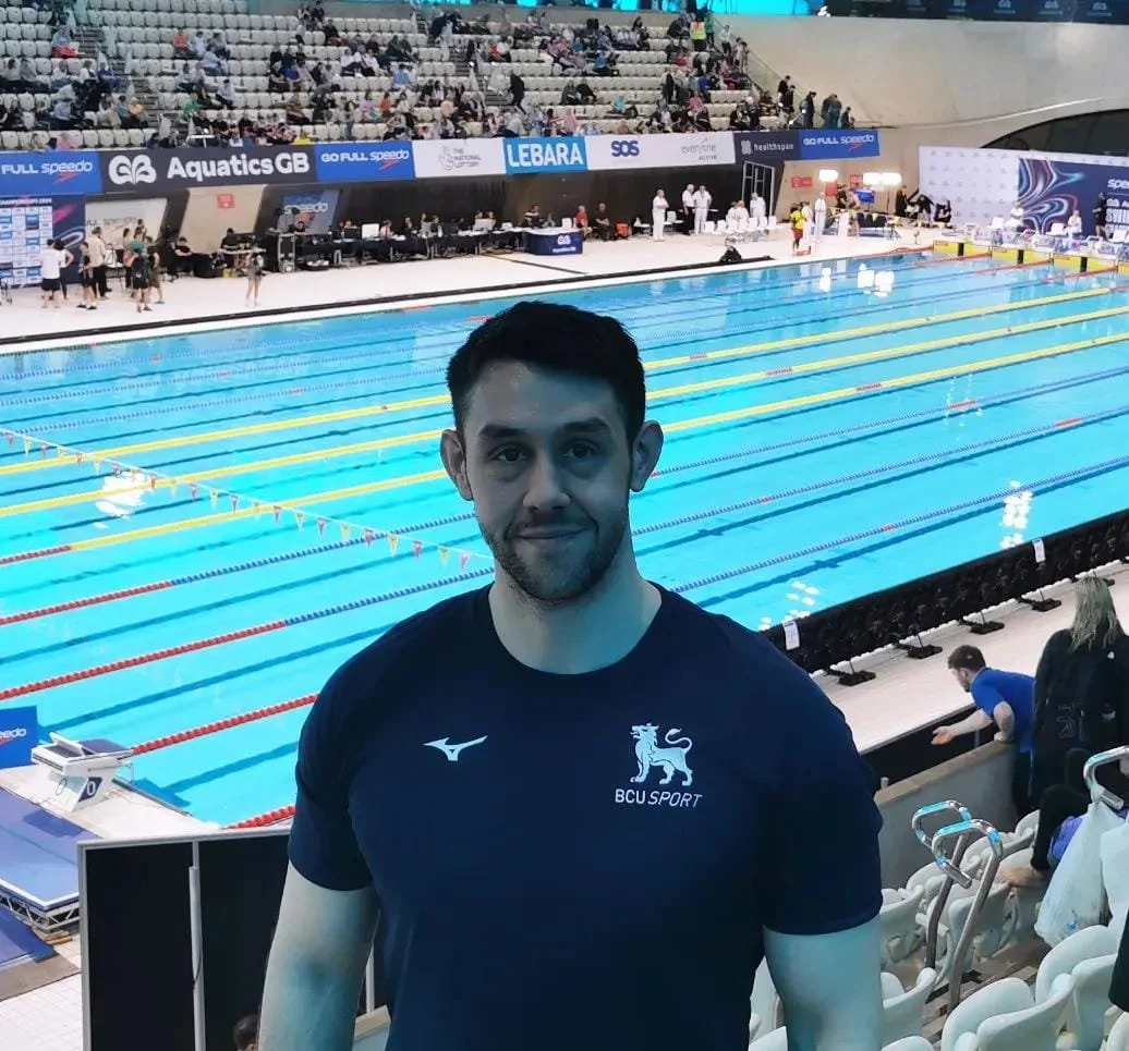 A man in a 'BCU Sport' shirt standing in front of an indoor swimming pool with lanes, surrounded by spectators in stands at a swimming event.