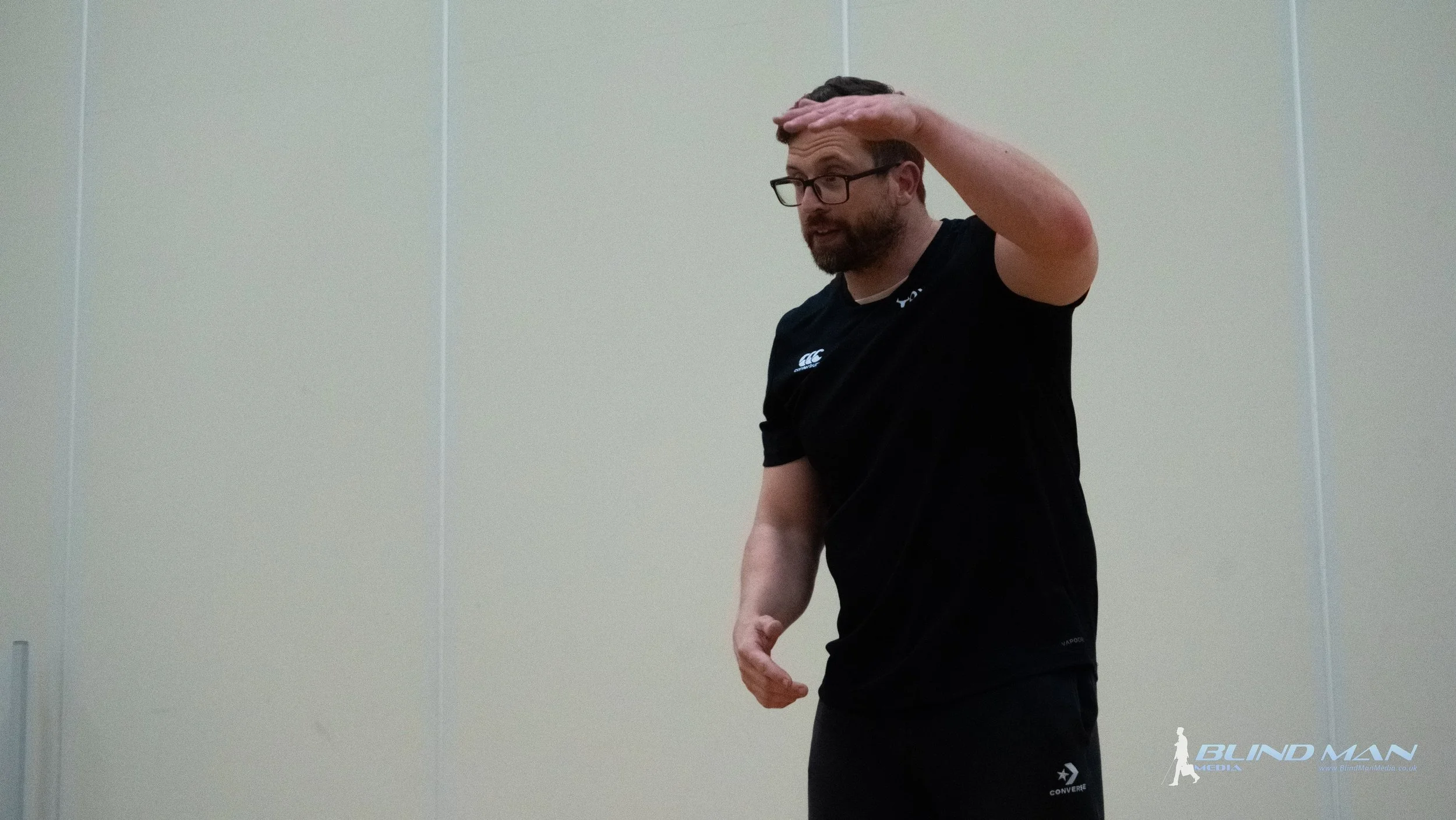 Man in a black shirt gesturing with his right arm raised in front of a beige wall.