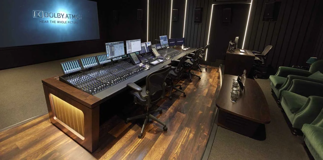 A professional audio and video editing studio with monitors, control panels, a large screen displaying a scene from a movie or TV show, and a comfortable seating area with a lamp.