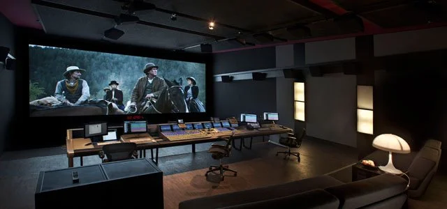 A professional audio and video editing studio with monitors, control panels, a large screen displaying a scene from a movie or TV show, and a comfortable seating area with a lamp.