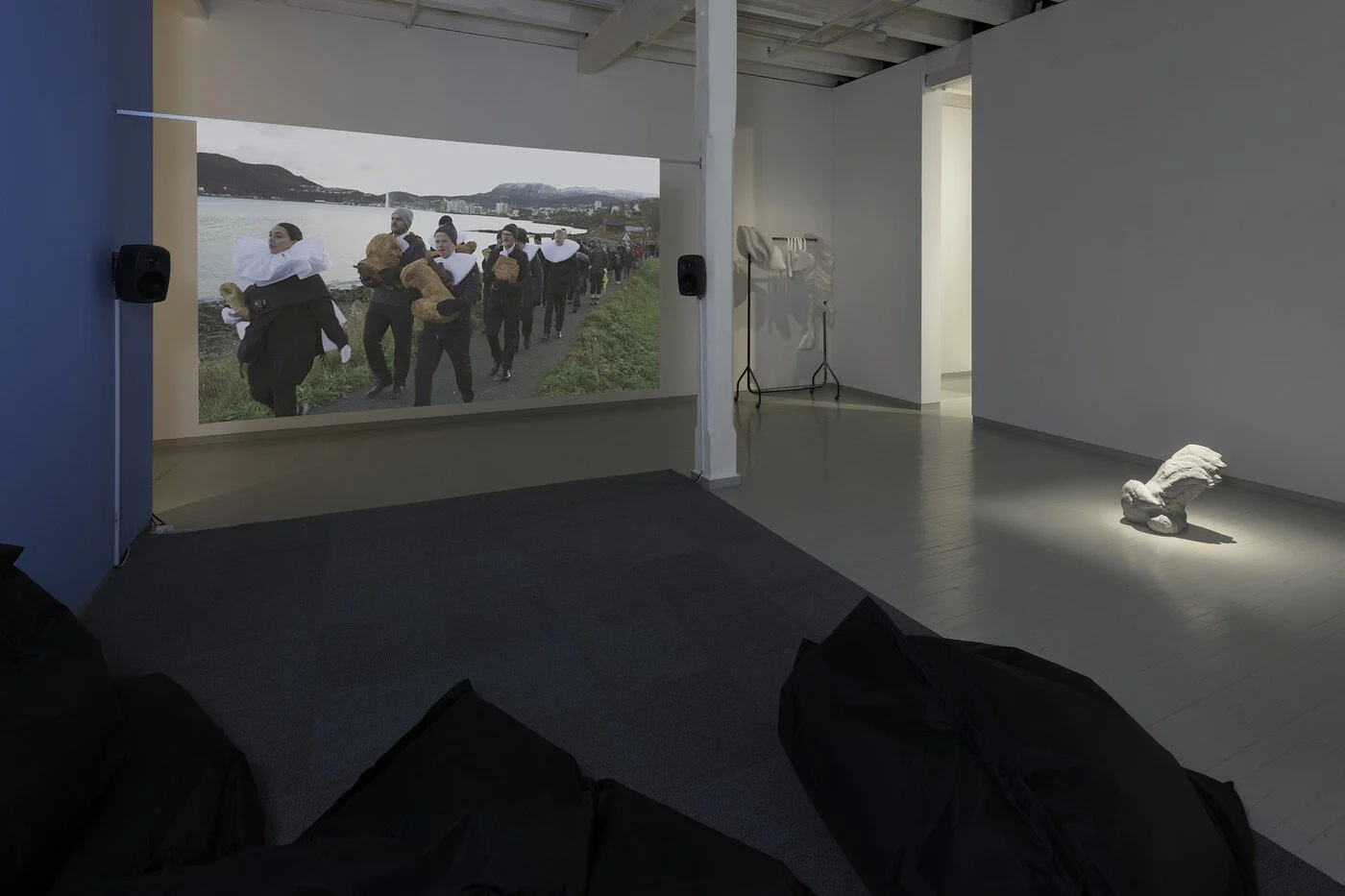 An art gallery exhibiting a large printed photograph of people carrying loaves of bread outdoors near a body of water. On the gallery floor, there is a sculpture of a folded cloth or blanket and a white sculpture of a reclining dog illuminated by a s