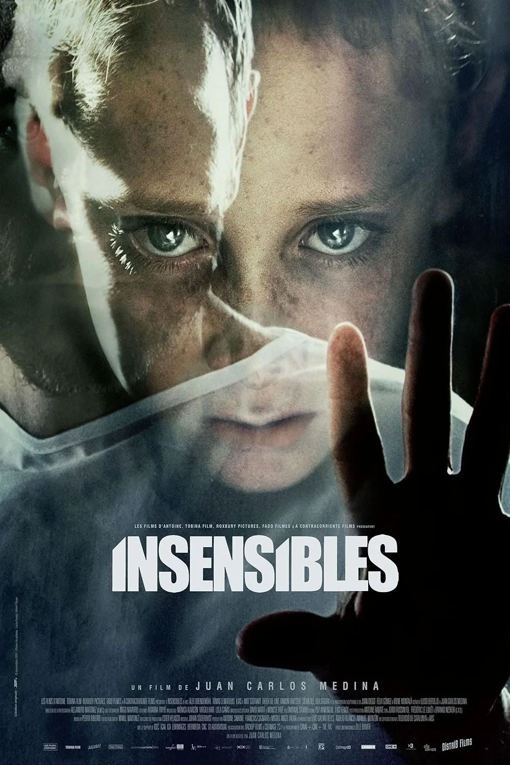 Close-up of a boy's face with intense eyes, wearing a clear face mask, creating a reflection of his face and hand pressed against a glass surface, with the title 'Insensibles' at the bottom.