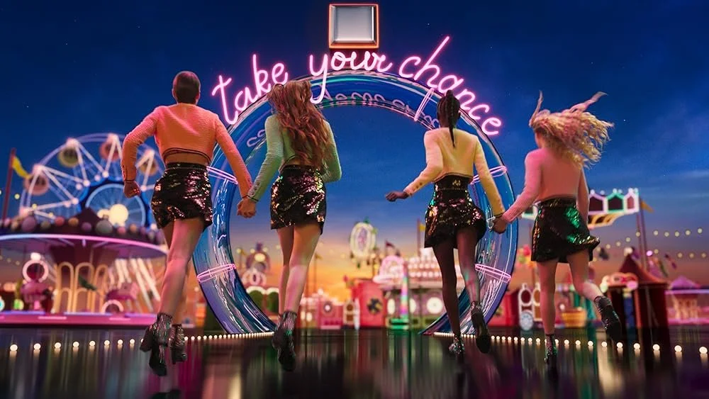 Four women in colorful outfits holding hands and jumping in front of a neon sign that says 'take your chance', with an amusement park in the background during sunset.