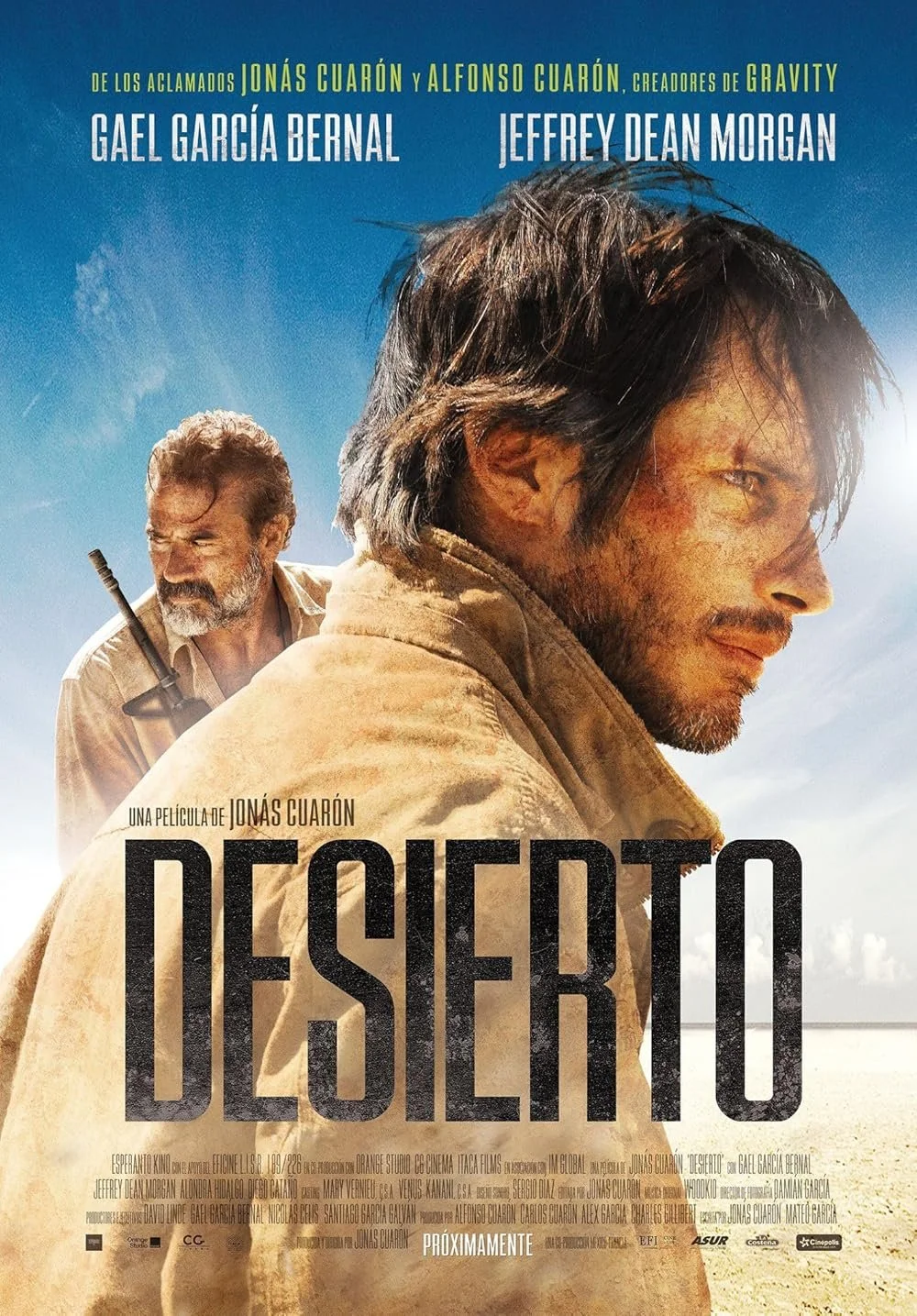 Movie poster for "Desierto" featuring two men in a desert landscape, one in the foreground with long hair and a sunburned face looking to the side, and another in the background with gray hair and a beard carrying a rifle. The sky is blue with some c