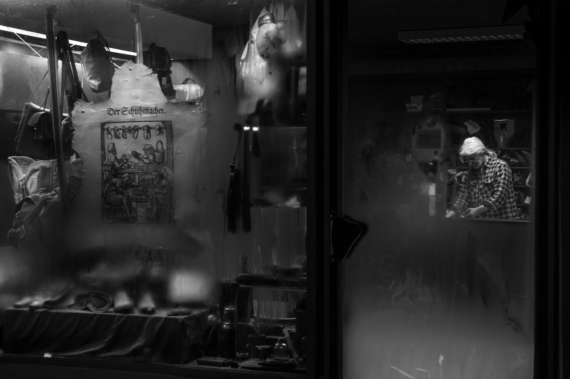 A black and white photo of a person with gray hair, wearing glasses and a checkered shirt, working in a workshop or store behind a glass window.
