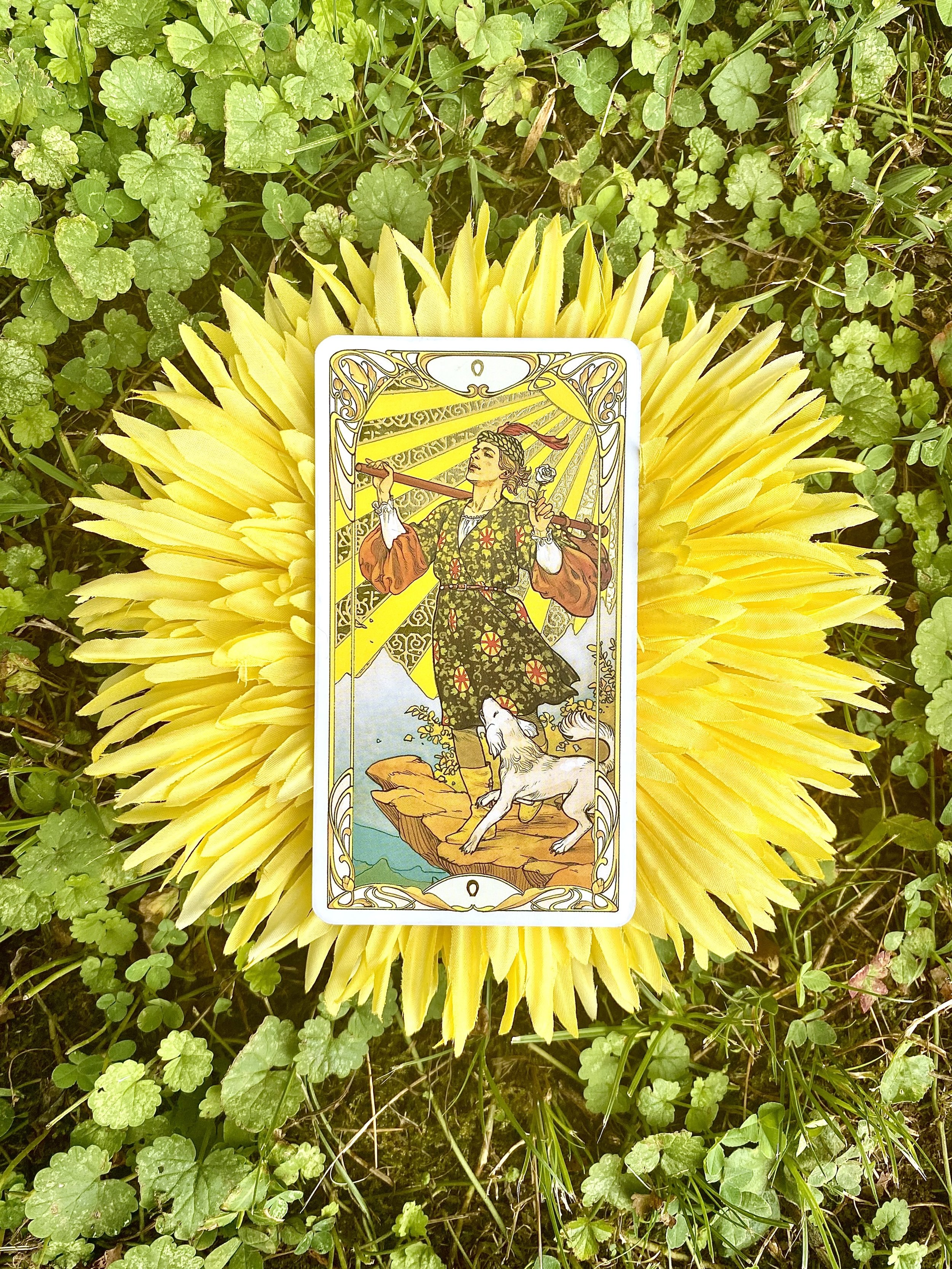 A tarot card featuring a woman in traditional folk attire with a dog at her feet, holding a rod across her shoulders with a sun and rays in the background, surrounded by green foliage and yellow flowers.