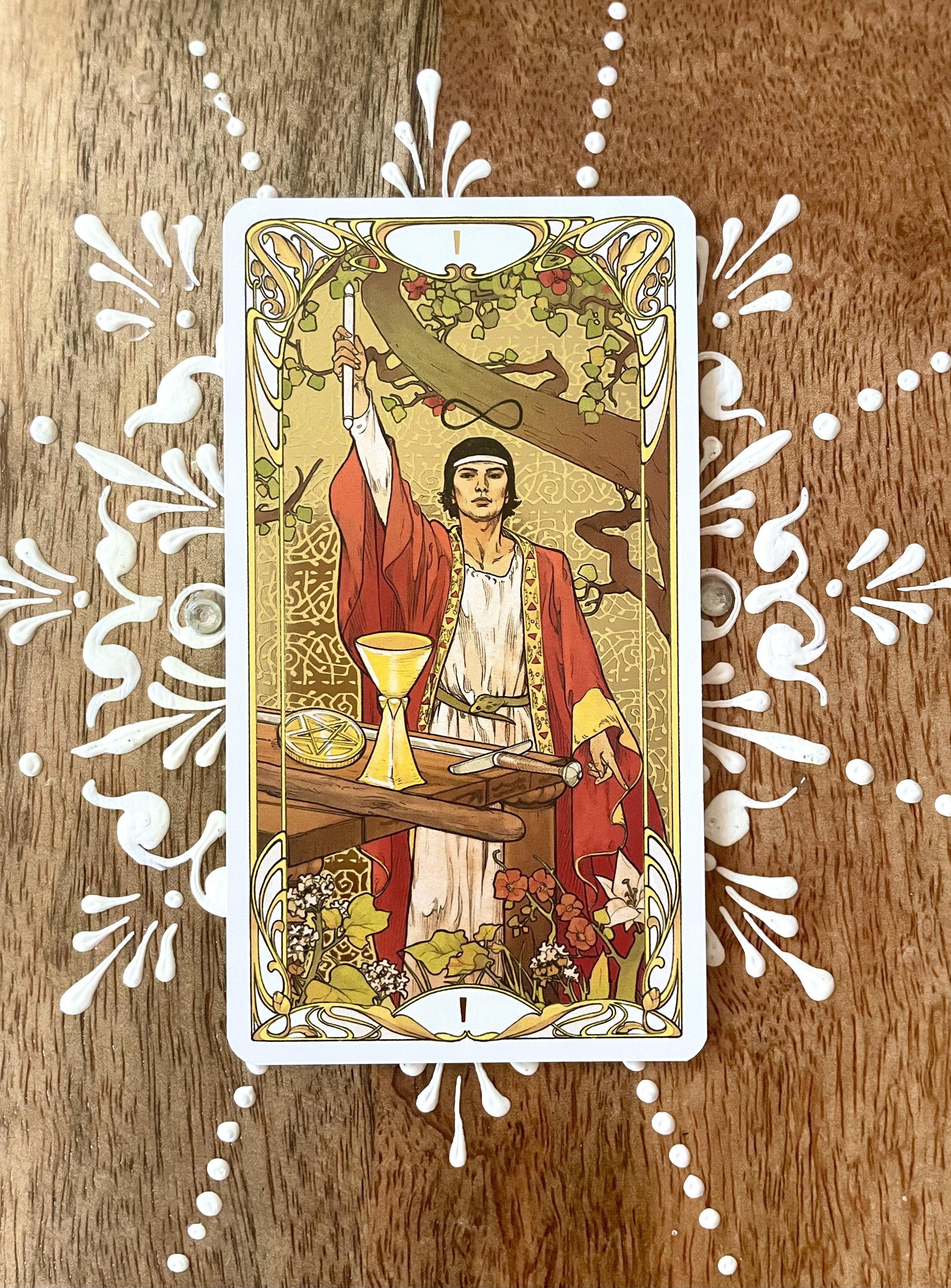 A tarot card featuring a person in a red robe holding a sword above their head, standing behind a table with a chalice and a sword, with a tree and floral background.
