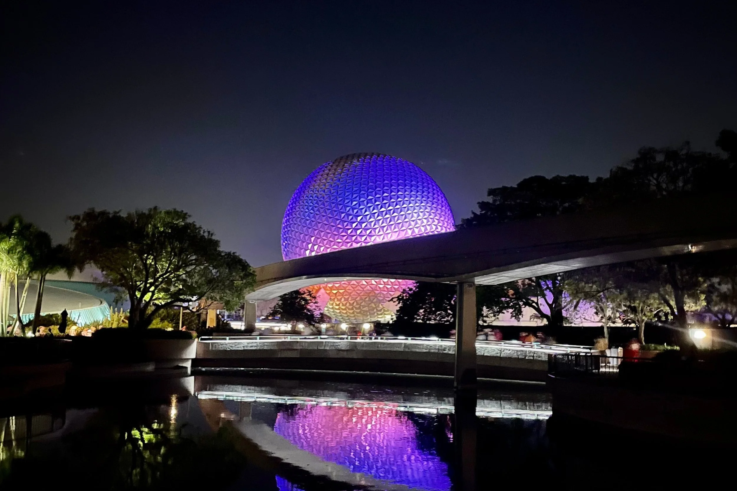 Epcot geodesic sphere illuminated with purple and pink lights at night, reflecting in a water feature in front, surrounded by trees and structures.