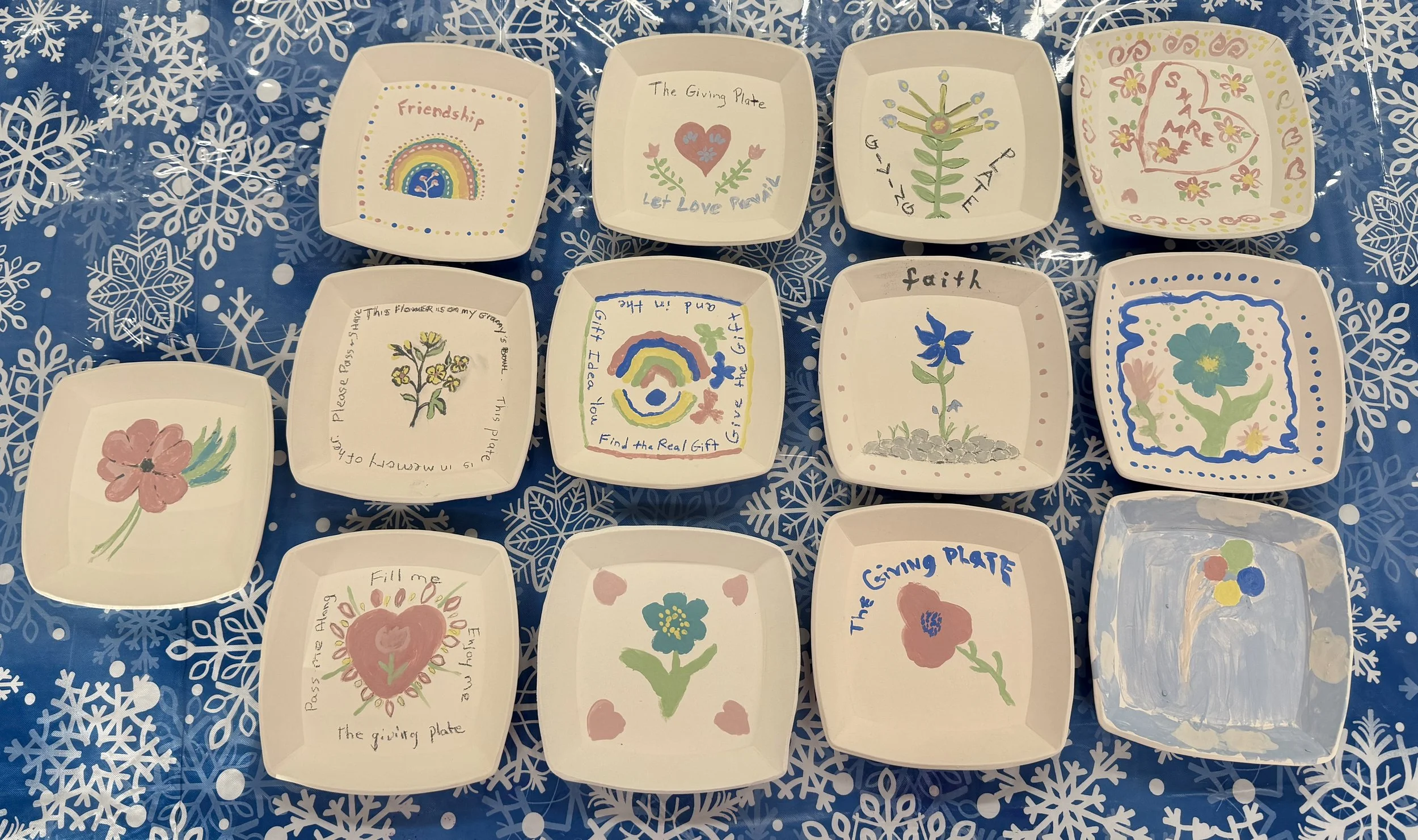 Collection of small decorated paper plates featuring colorful drawings and handwritten messages related to the themes of friendship, love, faith, and giving, arranged on a blue tablecloth with white snowflake patterns.