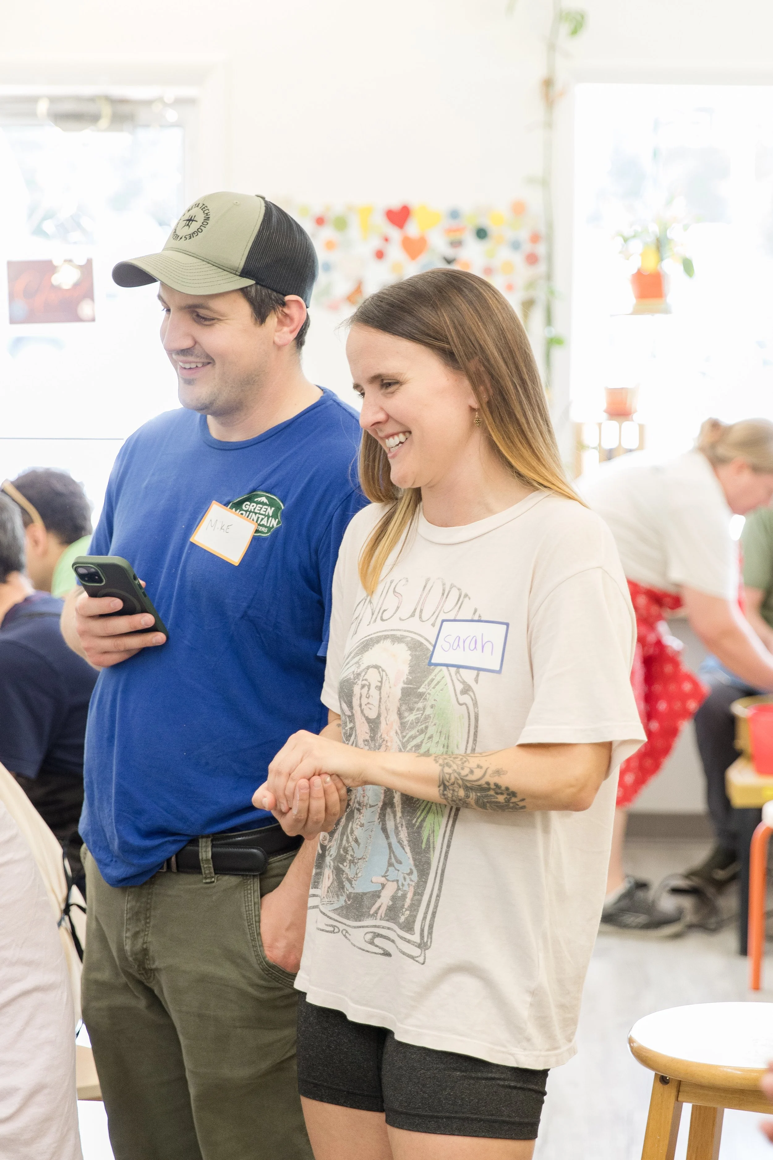 Two people standing indoors at a social gathering, smiling and looking at something off-camera. One person is wearing a blue shirt, a cap, and has a name tag that reads 'M'ke'; the other is wearing a white T-shirt with graphic art, a name tag that reads 'Sarah,' and has a visible tattoo on her left arm. They are in a decorated room with colorful hearts and dots on the wall, and large windows letting in natural light.