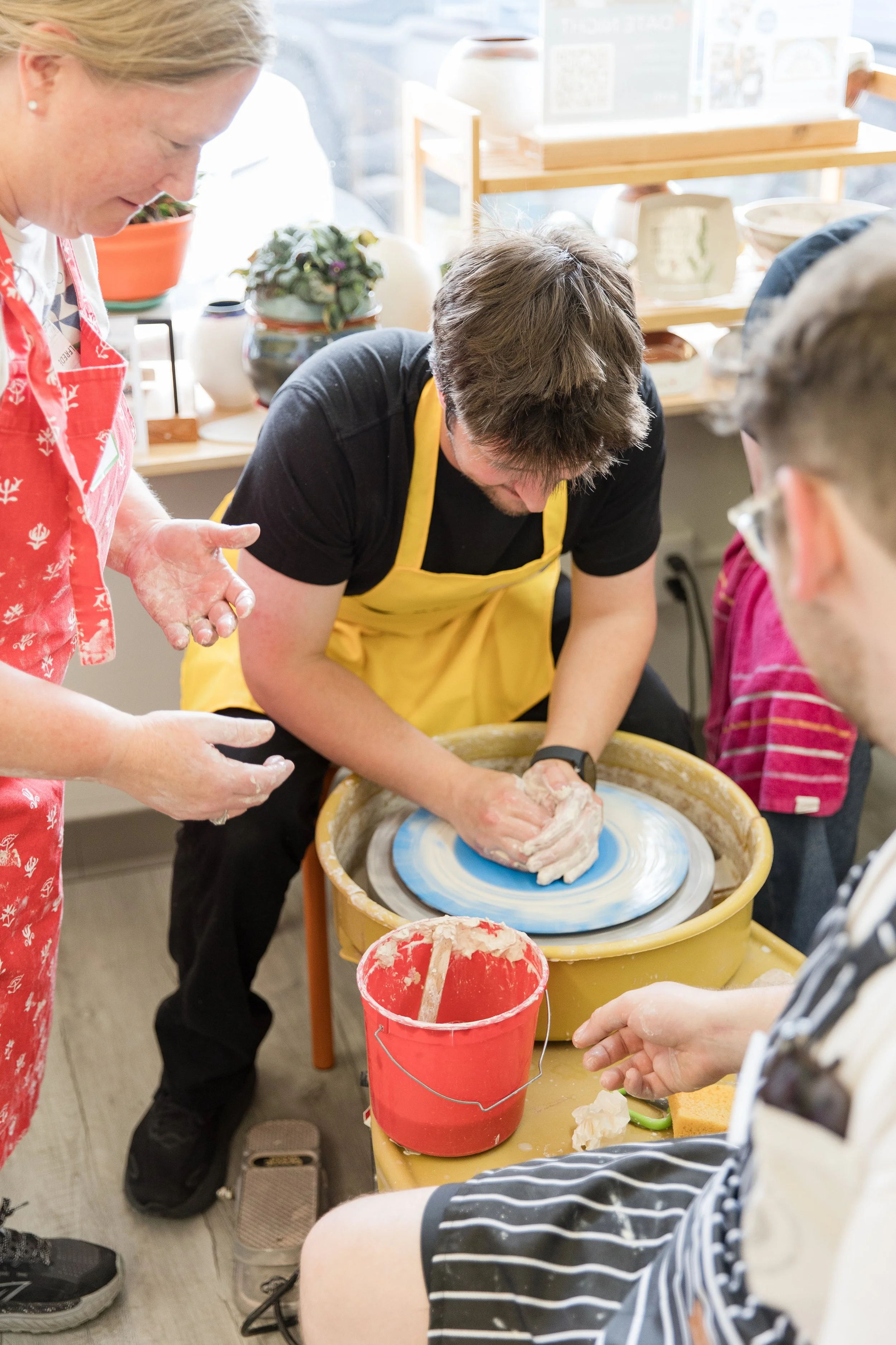 Group of people shaping clay on a pottery wheel in a pottery studio, with pottery tools and supplies nearby.