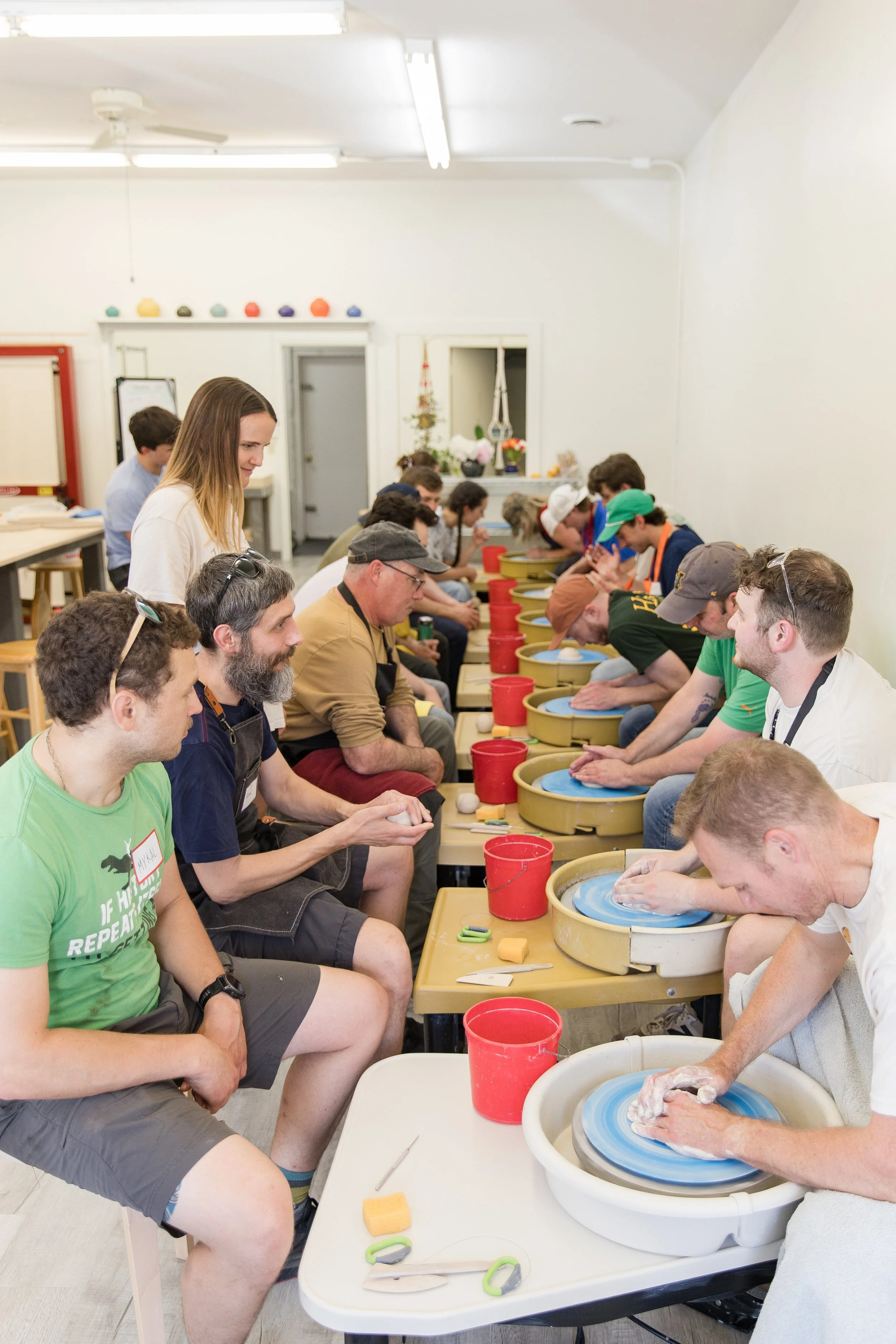 A group of people seated in a row at pottery wheels, making pottery items. Some are wearing aprons, and a female instructor is assisting a participant. The room is brightly lit with white walls, decorated with colorful balls and plants.