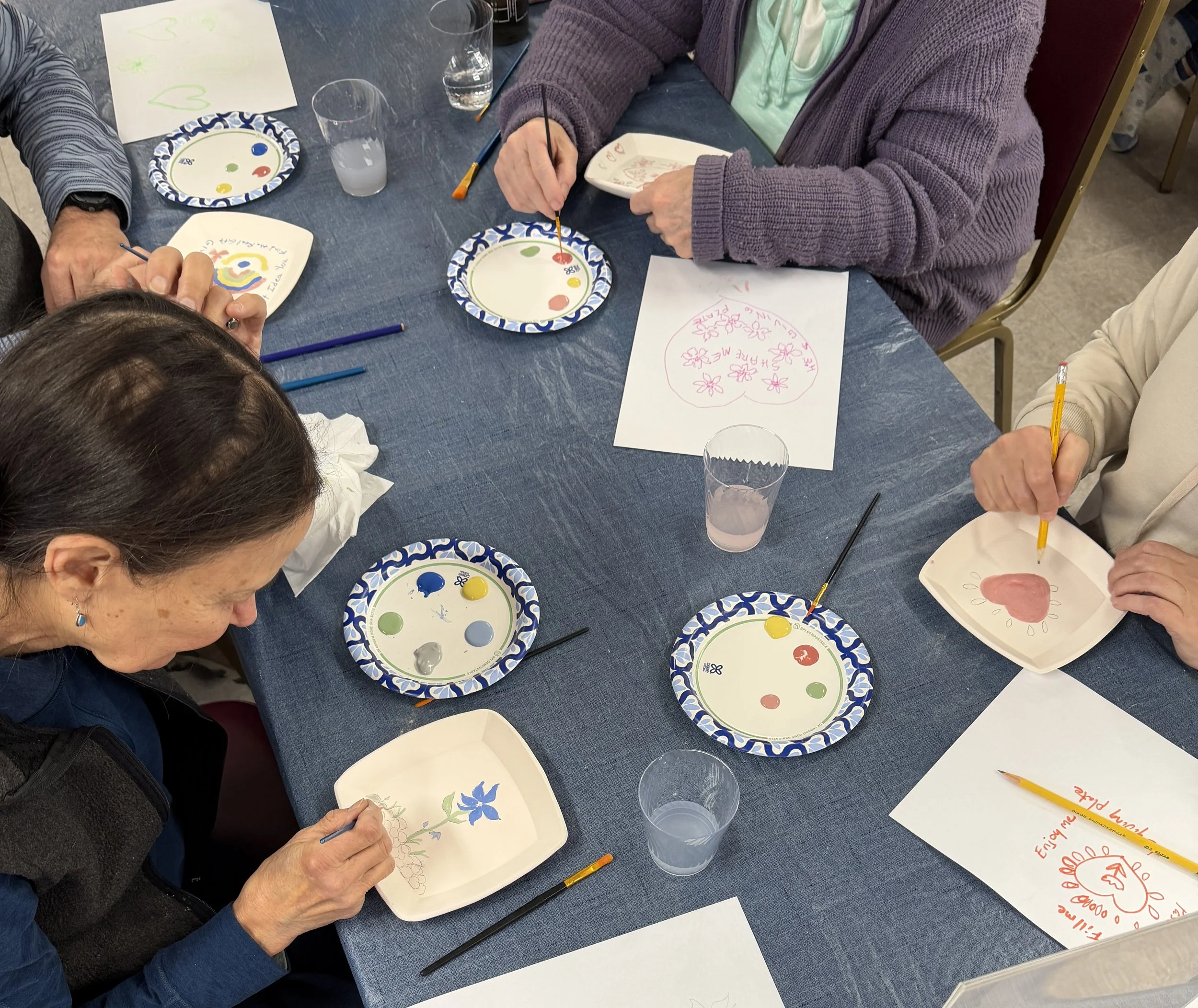 People sitting at a table painting and drawing on paper and plates with colorful paints.