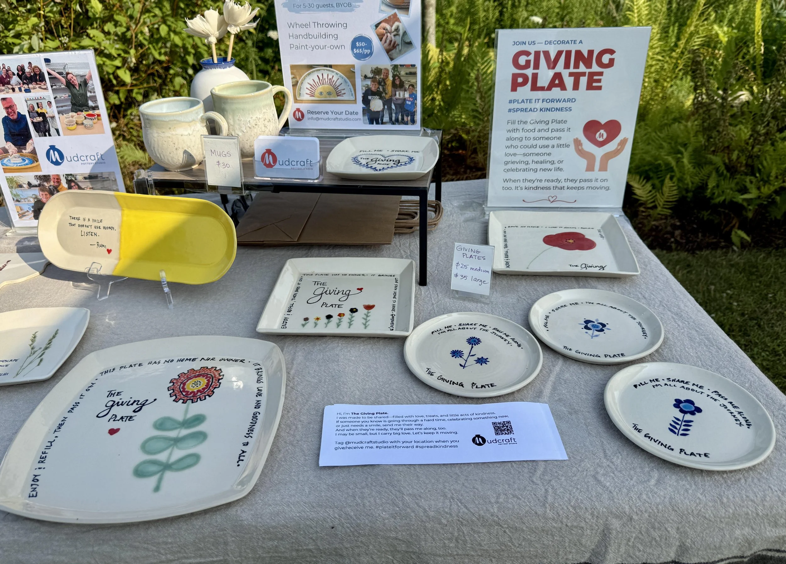 Table display of ceramic plates with inspirational messages and drawings, along with mugs, cups, and decorative items, at an outdoor event promoting giving and kindness.