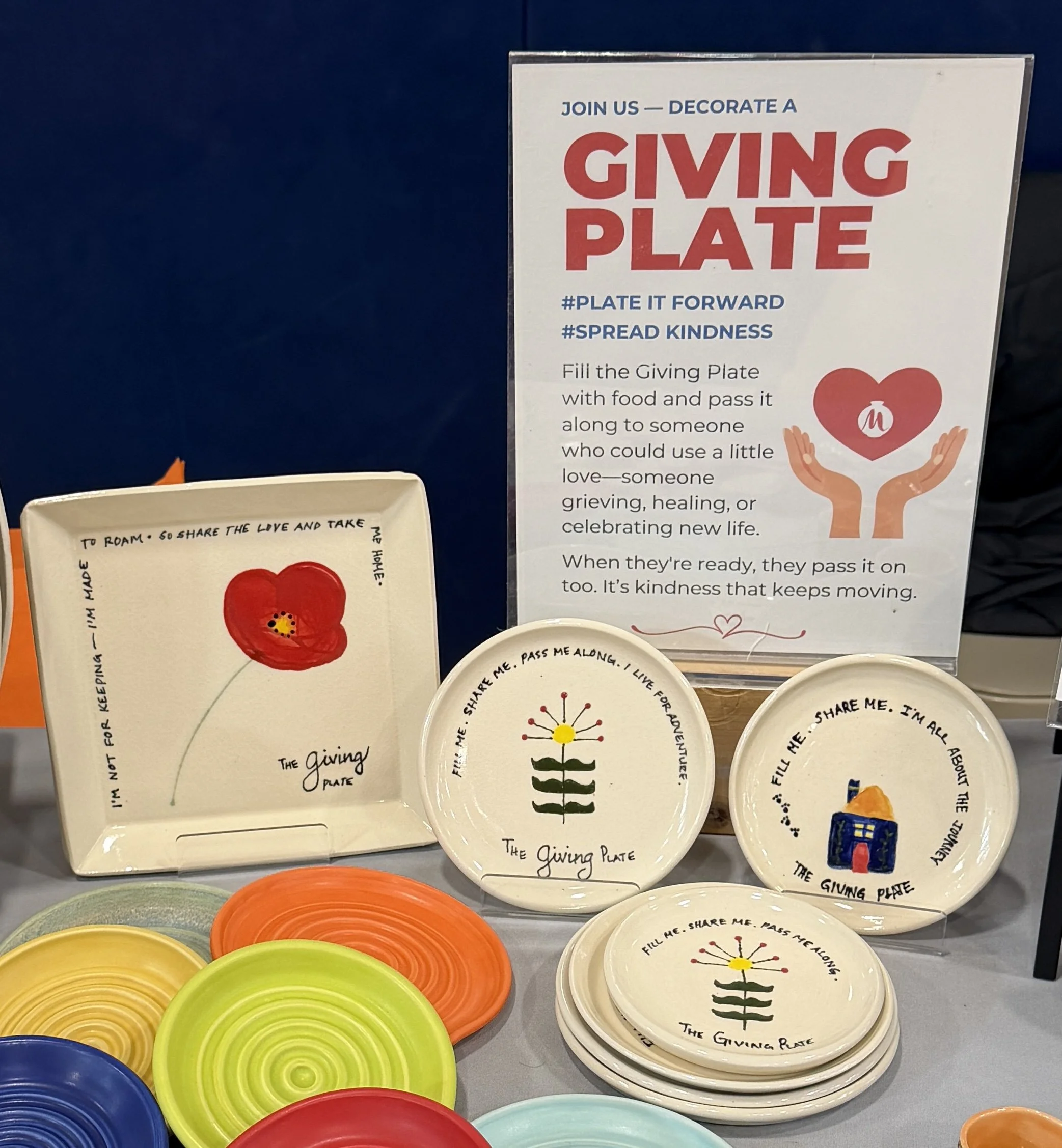Display of ceramic plates with colorful designs and a sign promoting a community giving activity. The sign encourages filling a plate with food to pass to someone in need, symbolizing kindness and sharing.