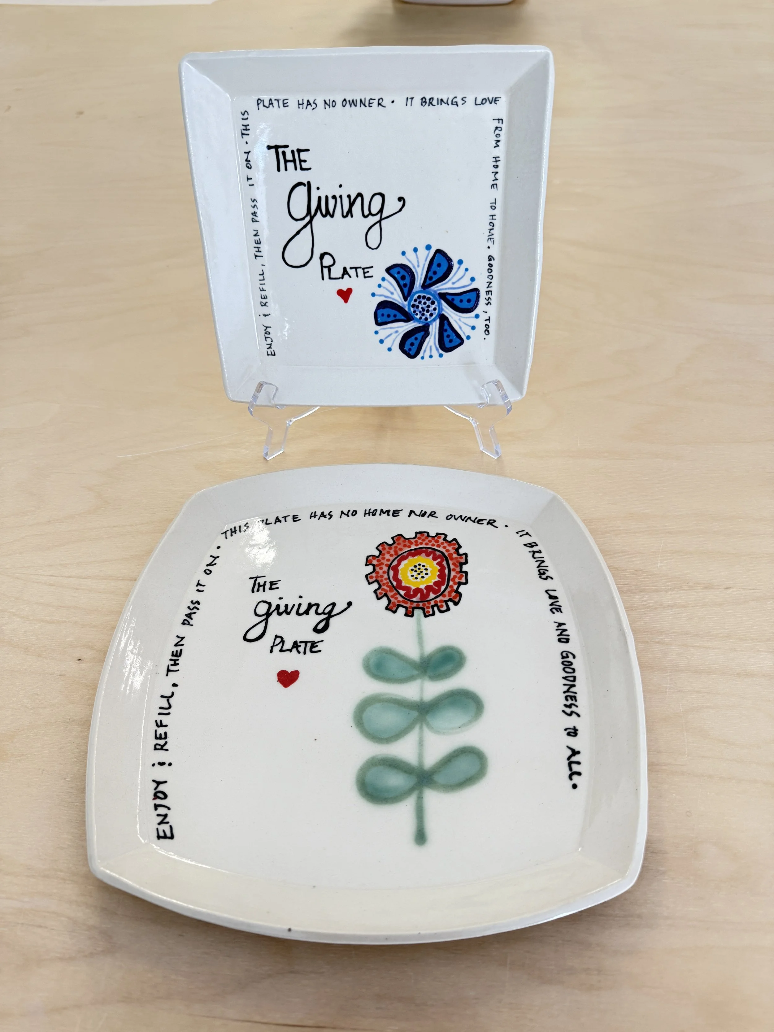 Two decorative plates with colorful hand-painted designs and inspiring quotes about giving, one placed on a stand and the other on a wooden surface.