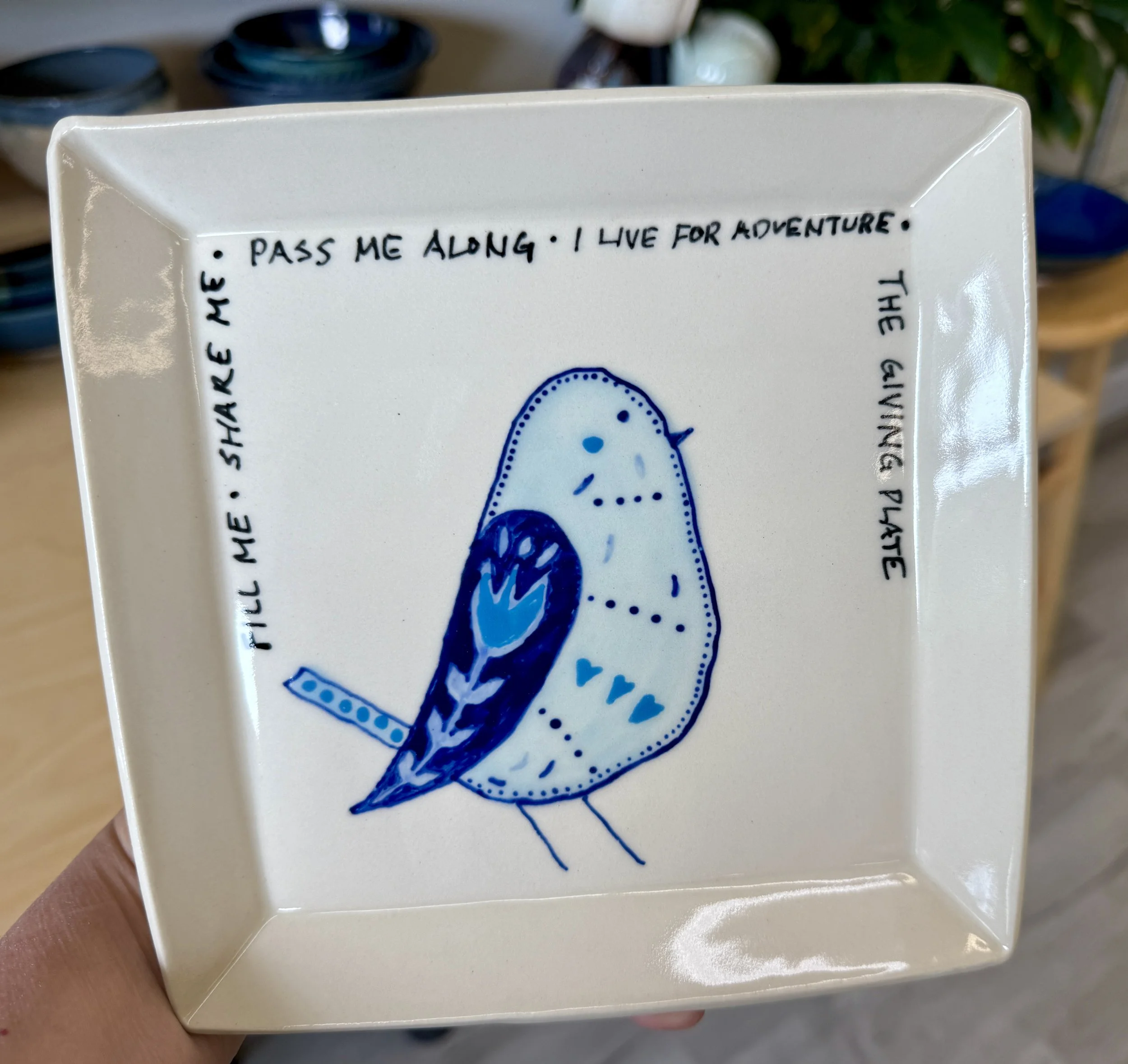 A decorative plate with hand-drawn art of a bird and a quote. The quote reads: "Pass me along. I live for adventure. The giving hand. Share me." The bird is illustrated in blue tones with simple lines and dots, standing on two thin legs.
