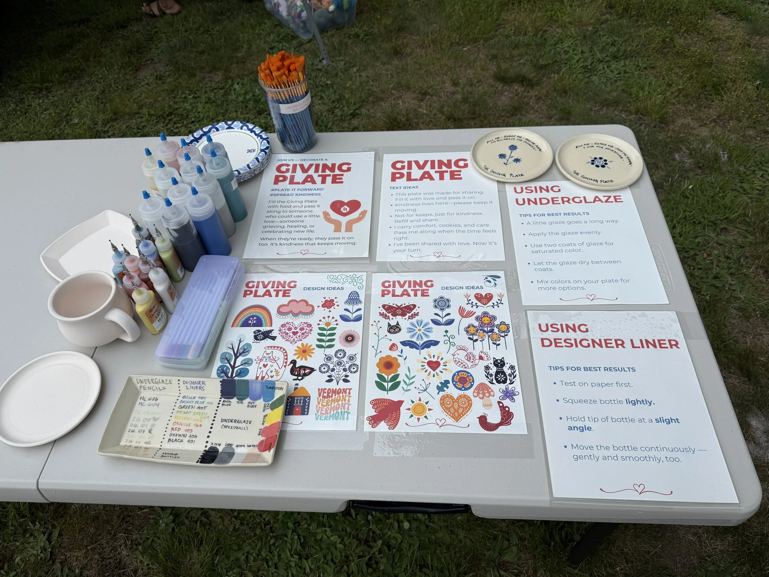 A white table set up outdoors with various supplies for creating decorative plates. There are bottles of paint, brushes, a cup, plates with printed instructions, colorful design sheets, and a container of underglaze labeled with color options. The table is on grass.