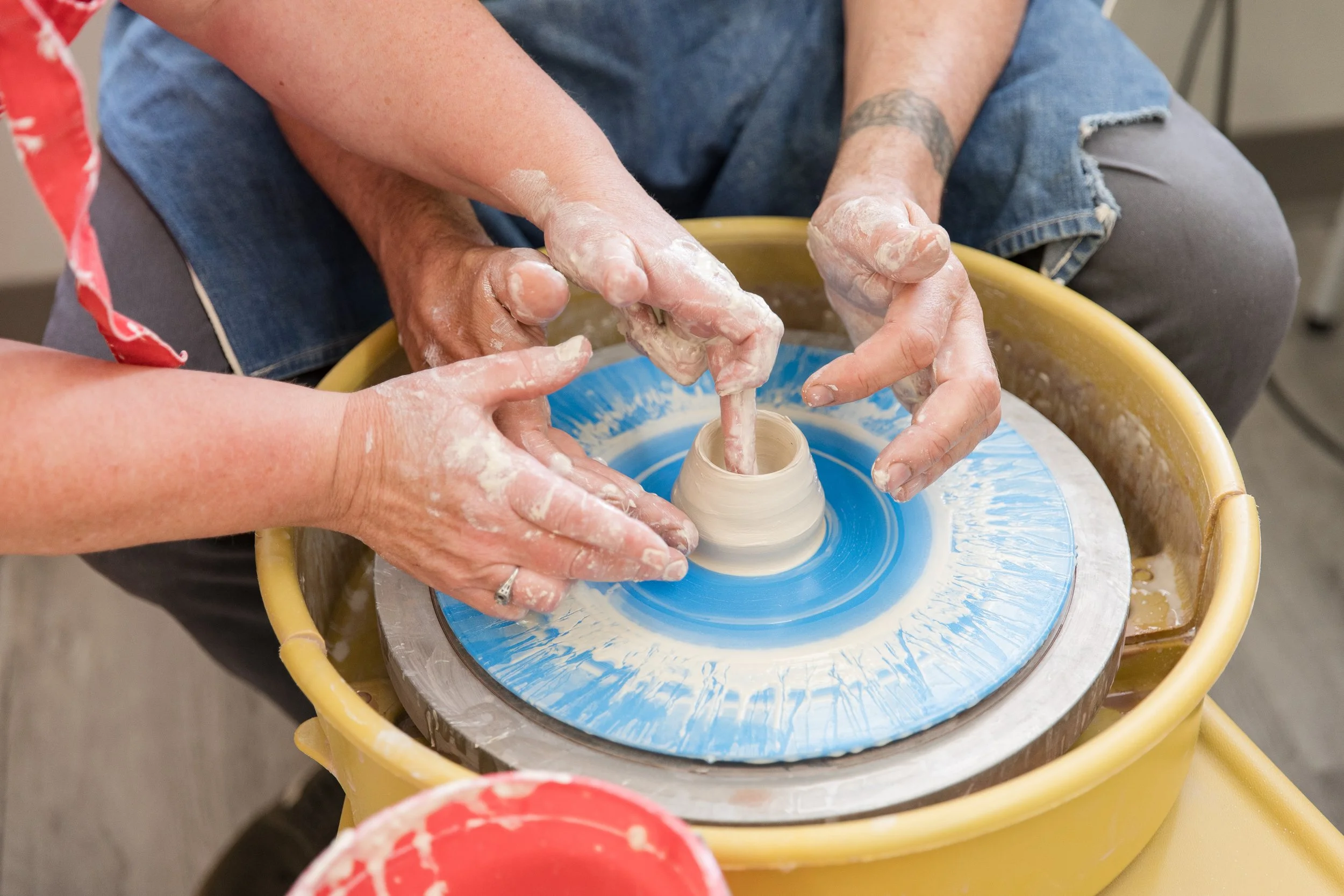 People shaping clay on a pottery wheel. A person with a tattooed arm is guiding the hands of another person as they work with the clay.