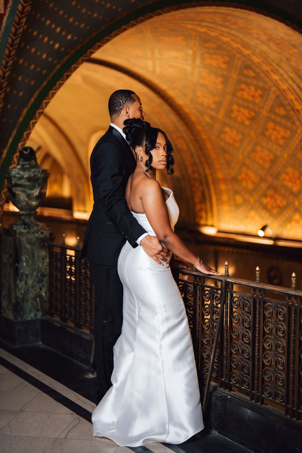 A couple dressed in formal attire, standing together on a balcony inside a grand, ornately decorated building with a rounded, patterned ceiling.