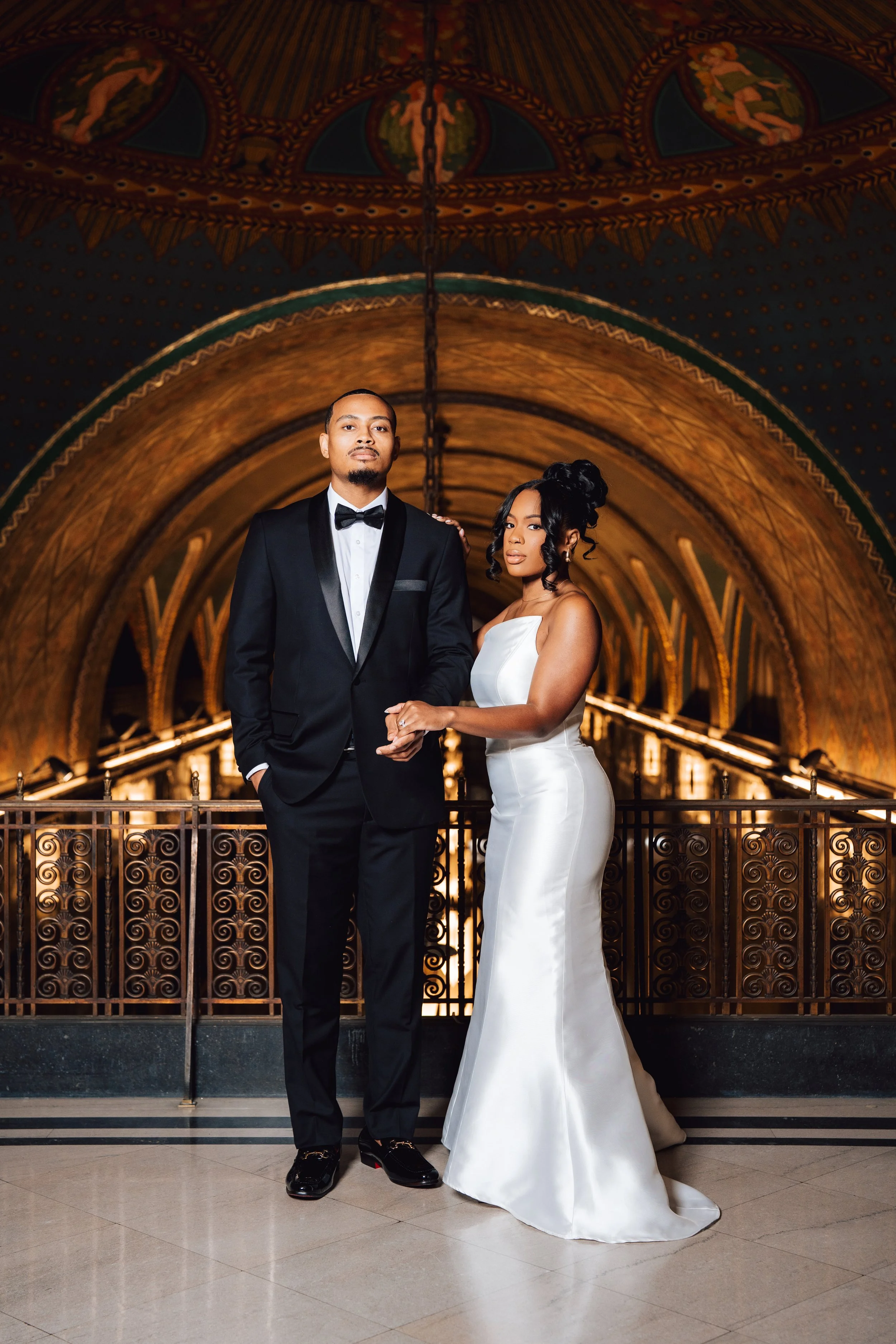 A couple in formal wedding attire standing in an ornate, historic building with gothic arches and gold detailing, holding hands and looking at the camera.