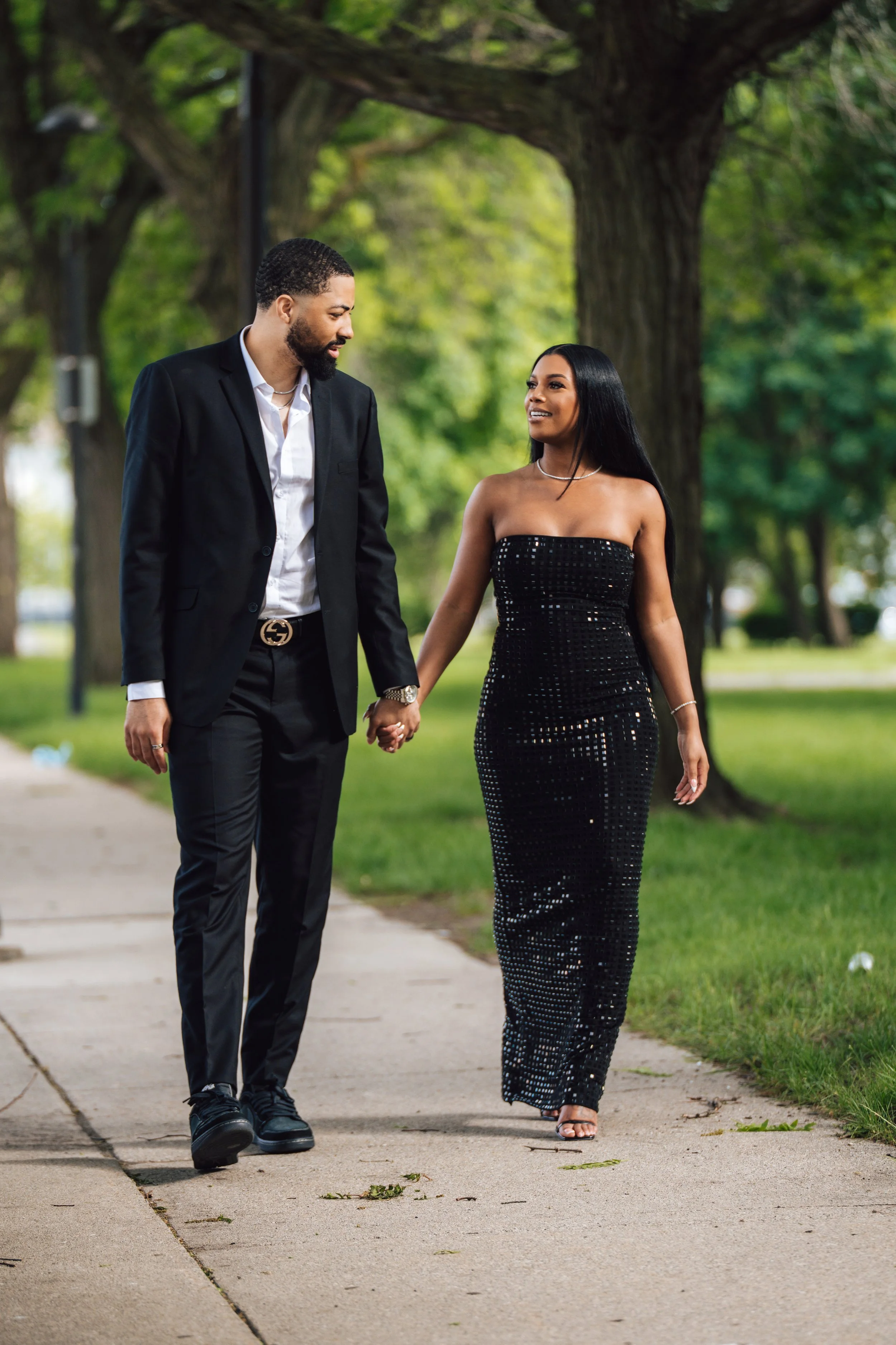 A couple holding hands walking on a sidewalk in a park, dressed in formal attire. The man is wearing a black suit with a white shirt, and the woman is wearing a strapless black sequined dress. They are smiling and looking at each other with trees and