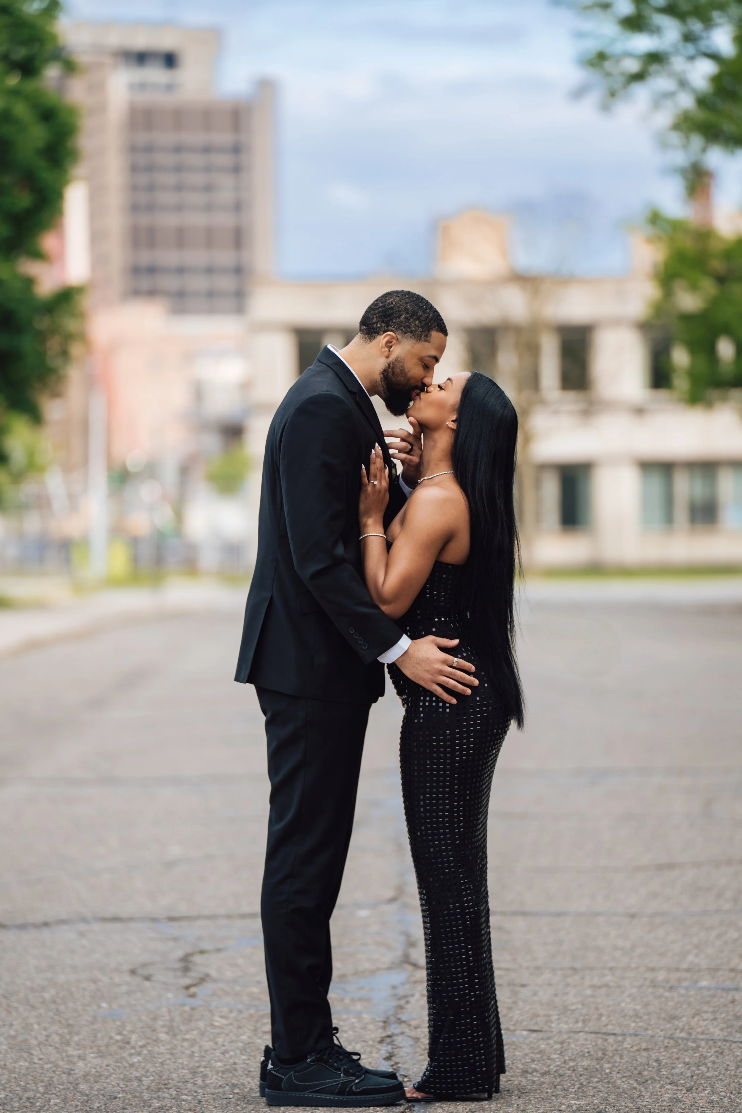 A couple dressed in formal attire sharing a kiss outdoors on a city street, with the man in a black suit and the woman in a black, dotted gown, surrounded by urban buildings and green trees.
