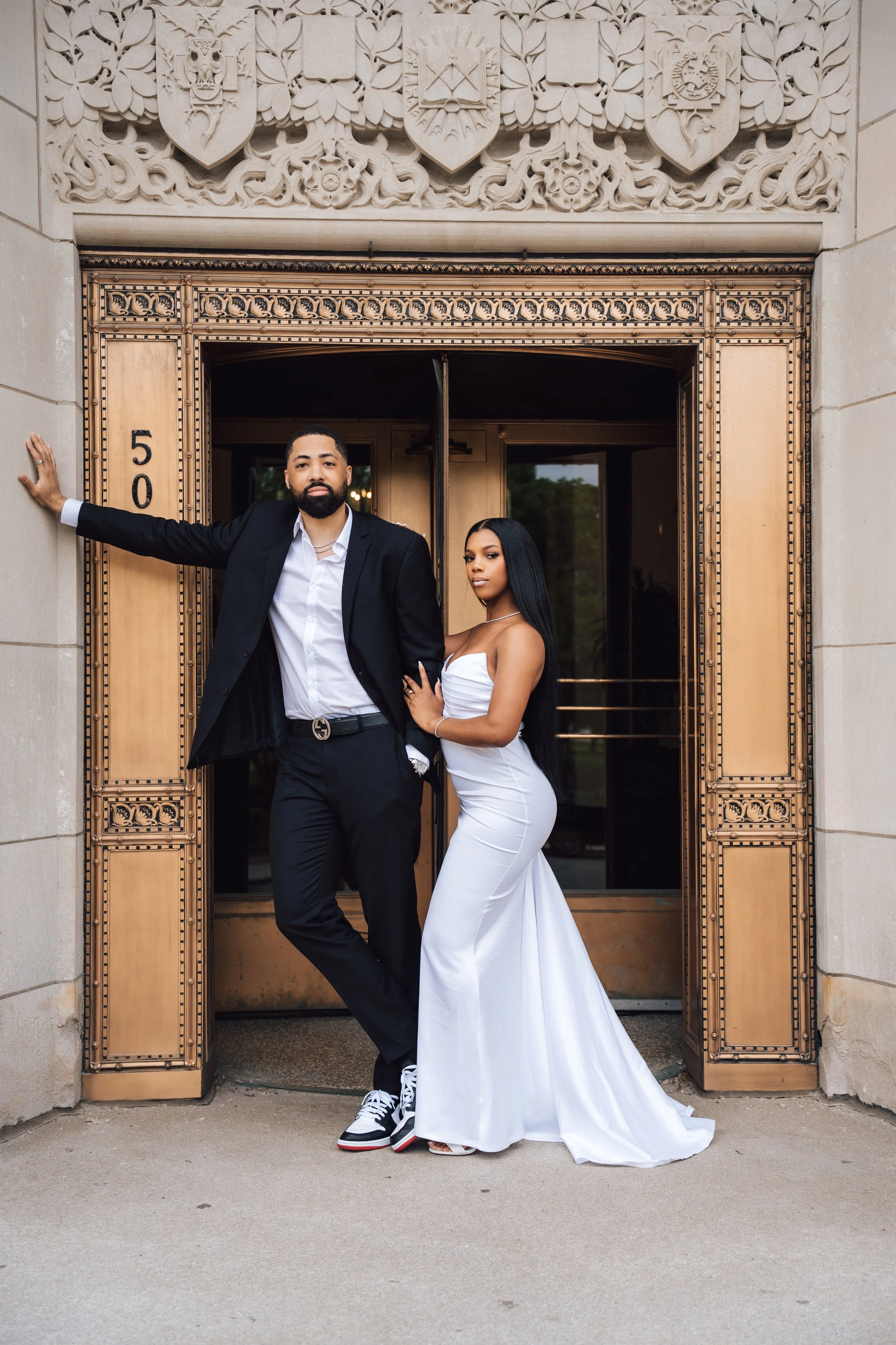 A stylishly dressed couple stands in front of an ornate building entrance with decorative carvings and shields. The man is wearing a black suit with a white shirt and sneakers, leaning against the wall. The woman is in a white strapless gown, standin