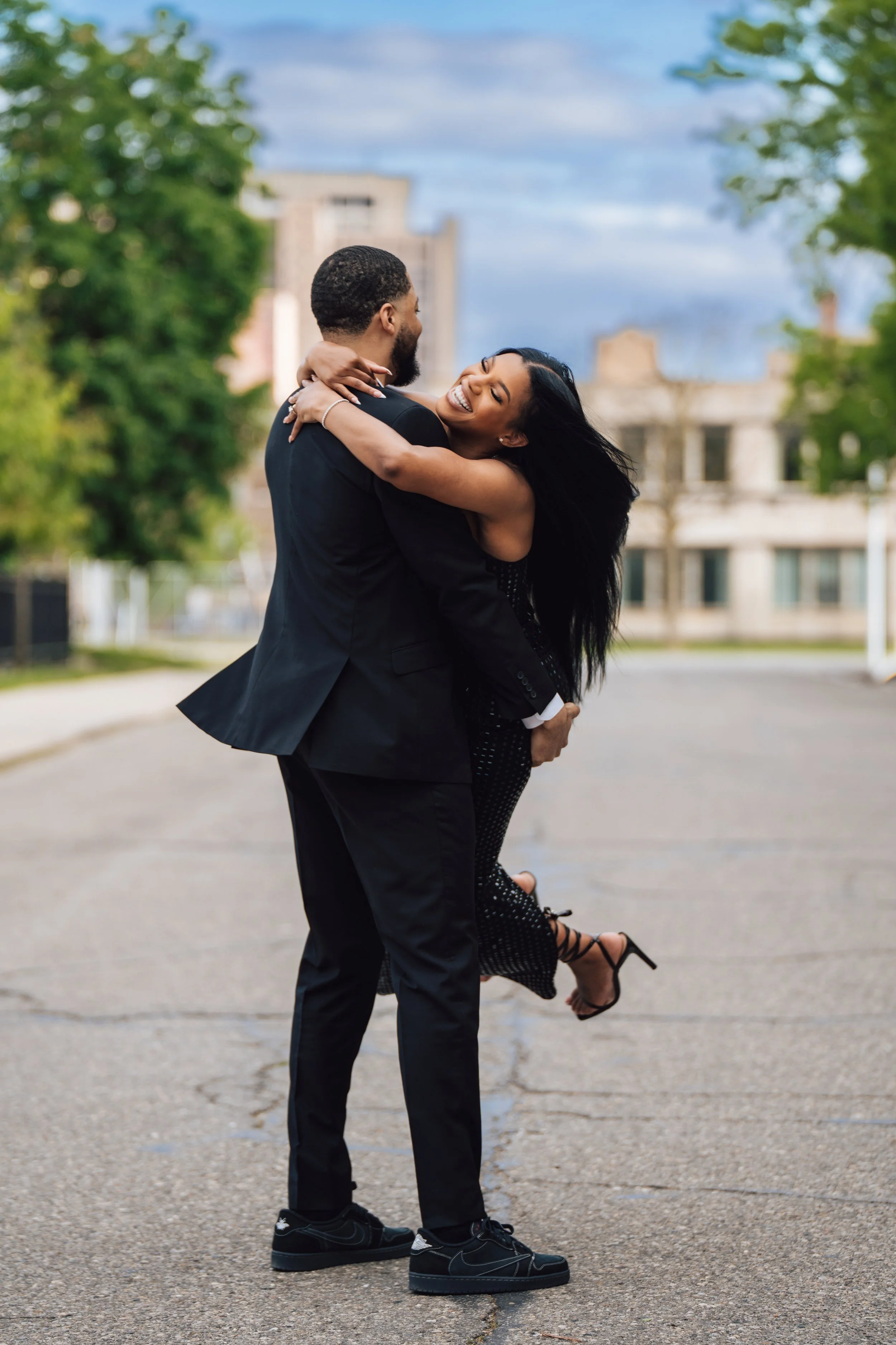 A man in a black suit lifting a woman in a black dress and high heels, both smiling and embracing outdoors on a paved surface with greenery and buildings in the background.
