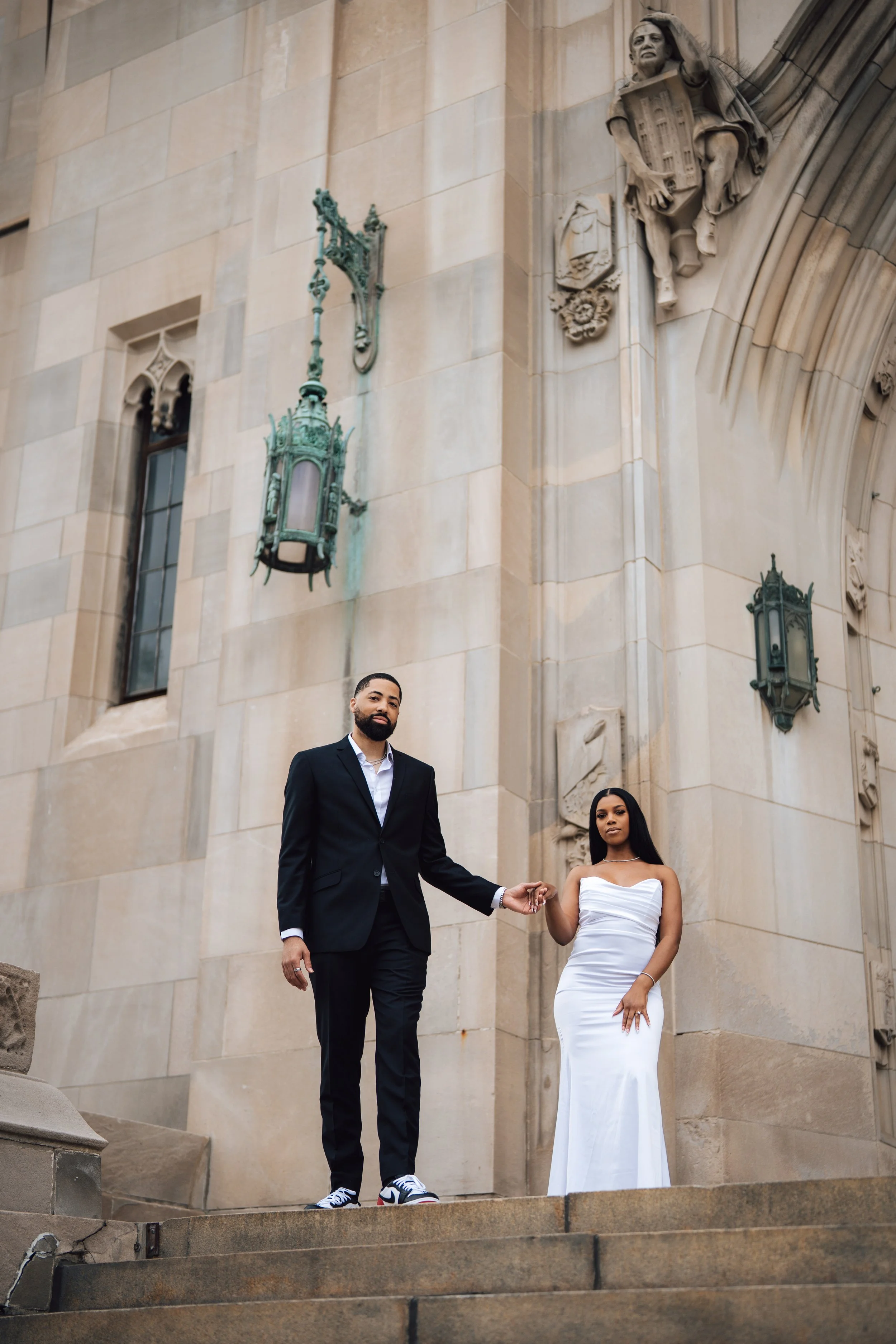 A man and woman in formal attire standing on steps outside a historic building with ornate architectural details and sculptures, holding hands.