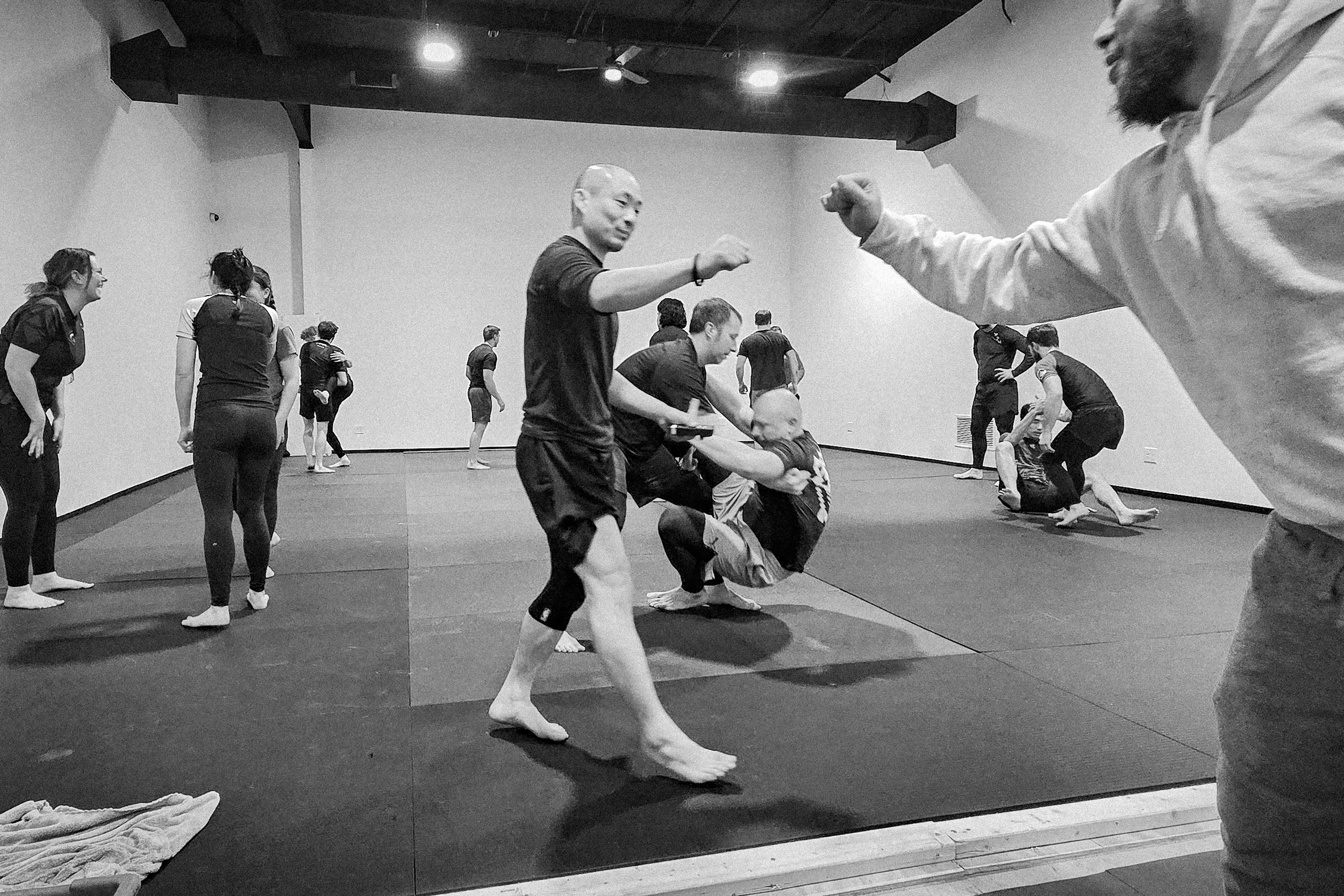 People practicing Brazilian Jiu-Jitsu in a martial arts training room with mats, some are sparring, others observing or waiting.