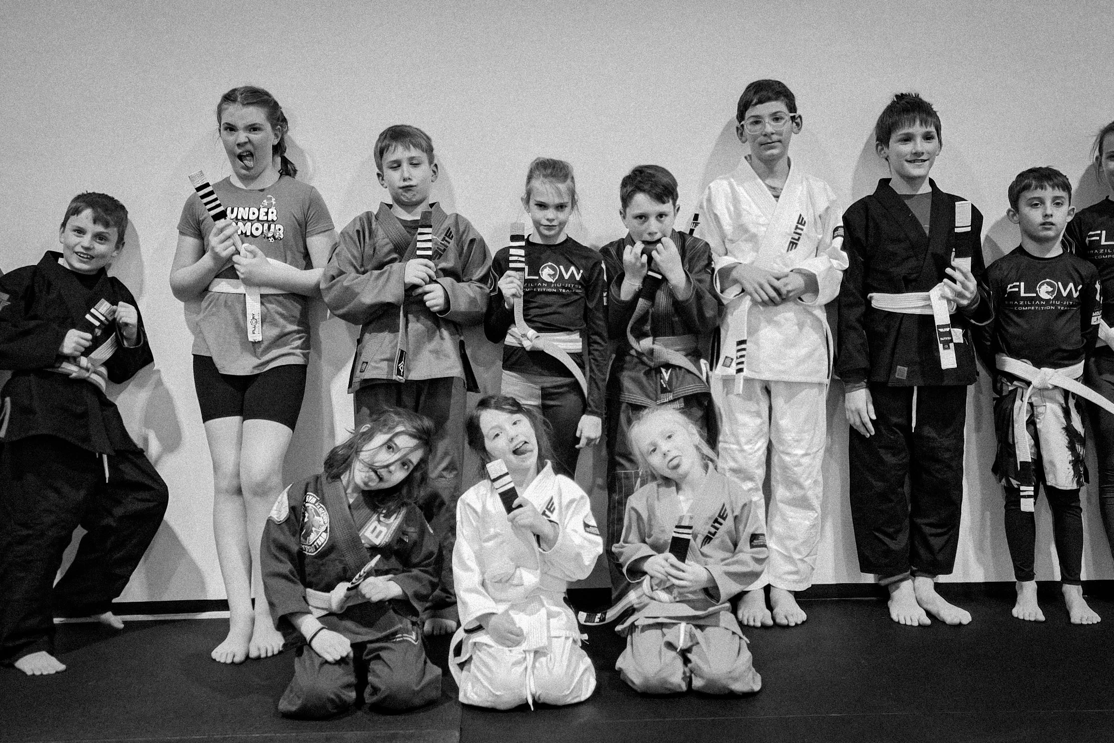 Group of children in martial arts gi and uniforms posing with belts after a class, some making playful faces.