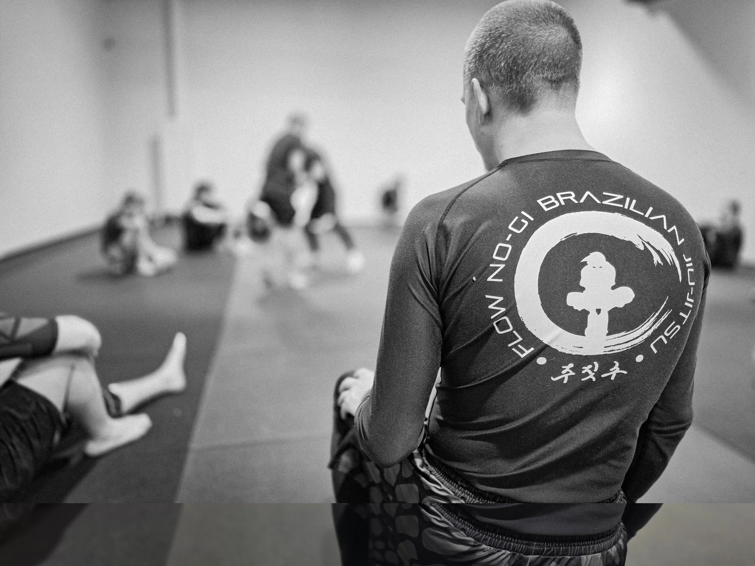 A male martial arts instructor in a Flow Brazilian Jiu Jitsu shirt observing a class with students sitting on the mat in training.