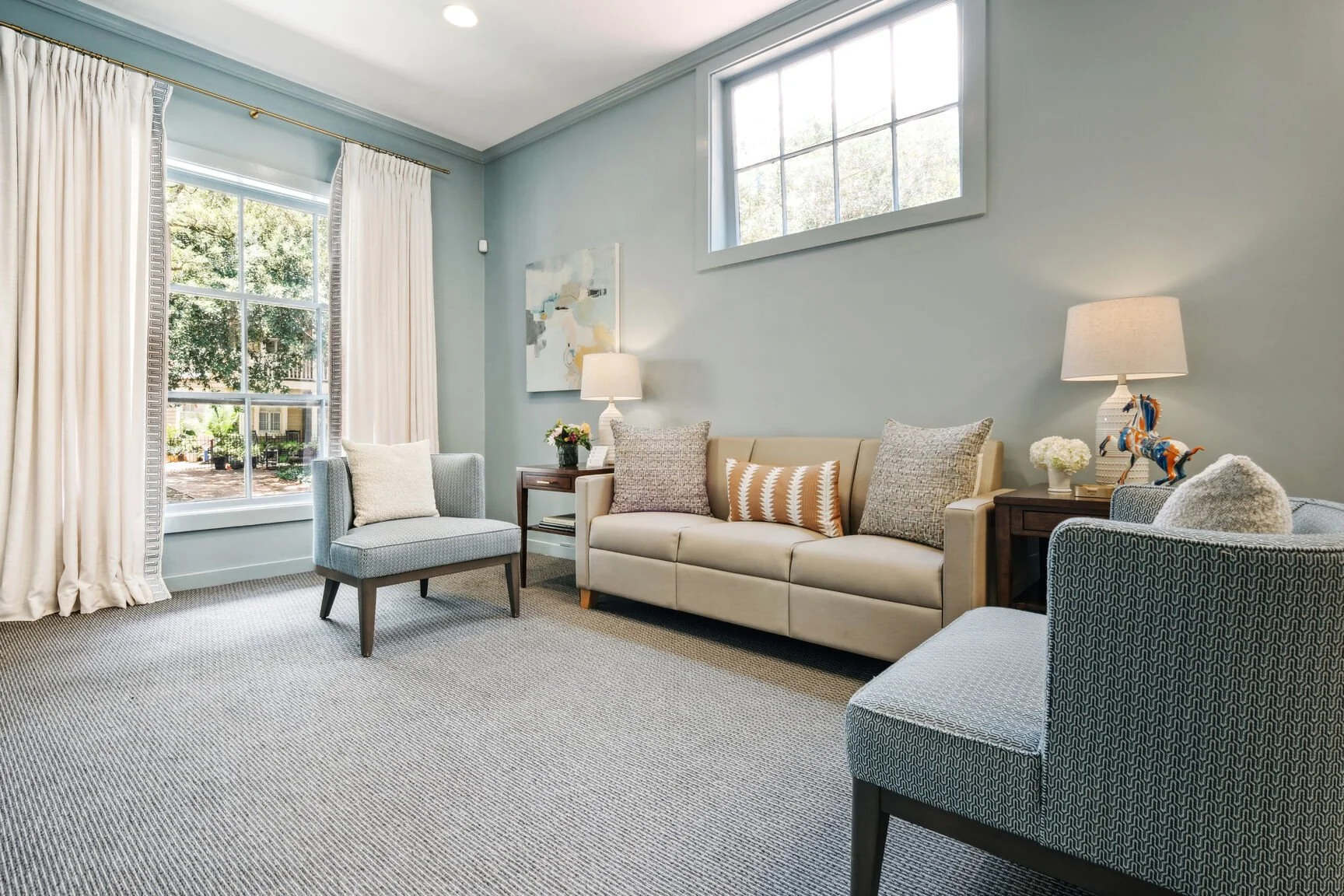 Bright living room with large windows, light blue walls, a beige sofa with patterned pillows, two grey armchairs, side tables with lamps, and wall art, decorated in neutral tones.
