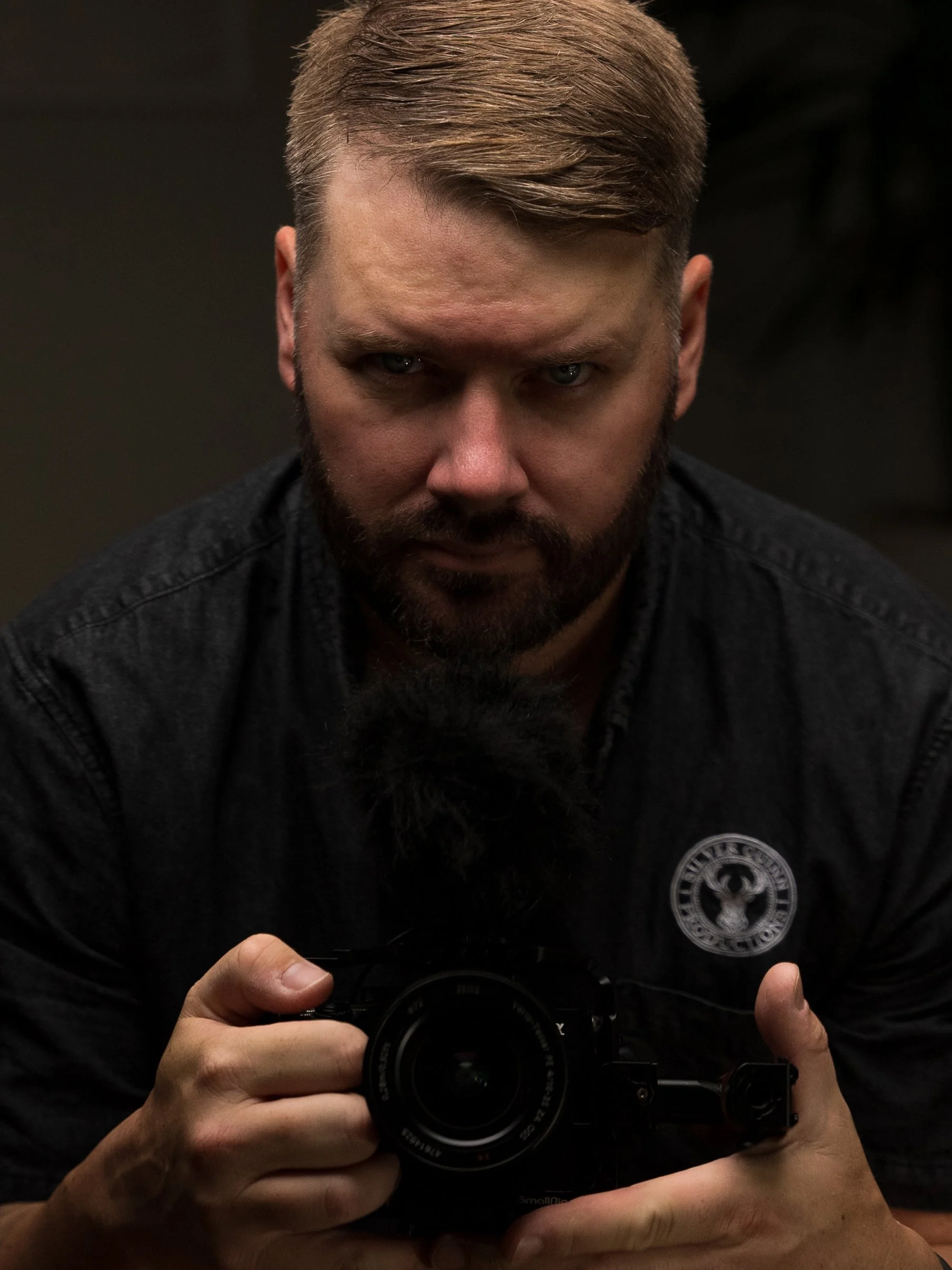 Man in a suit with a deer pin, dark hair and beard, looking at the camera.