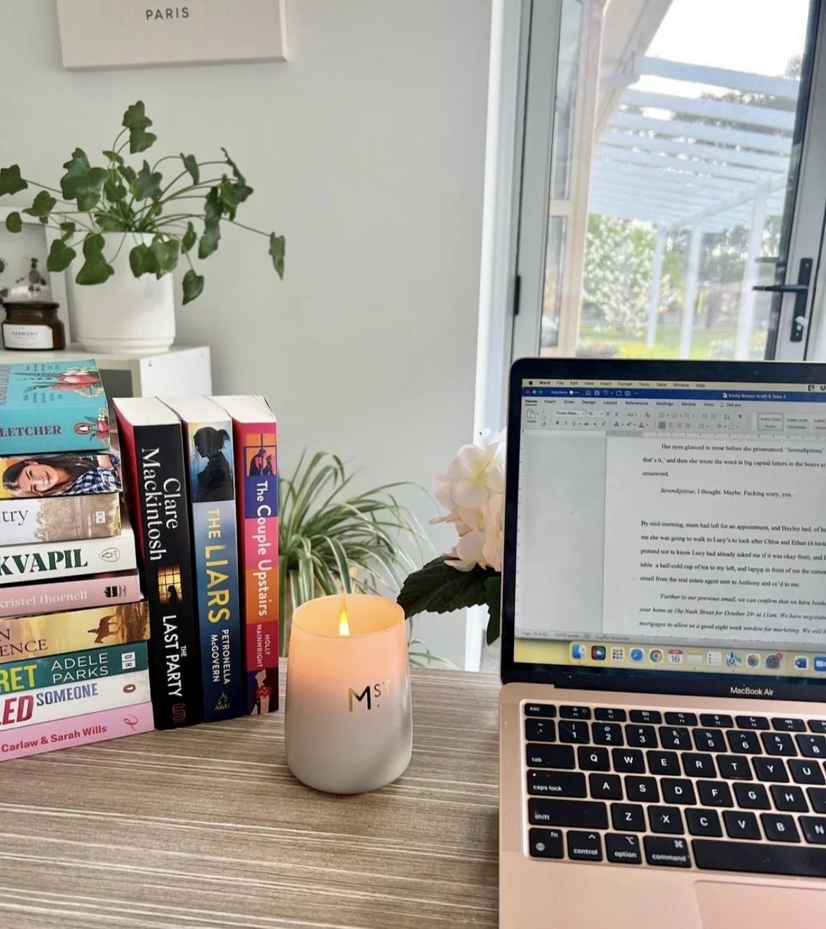 Desk with books, laptop and candle. Pot plant behind in the distance.