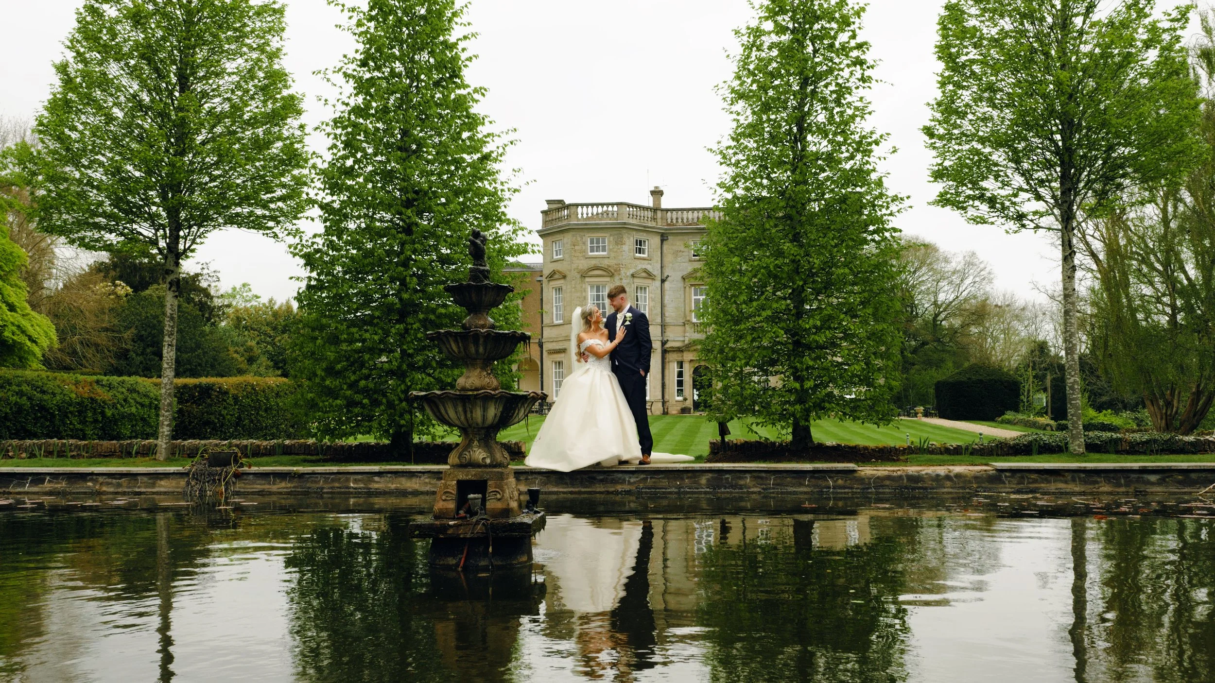 The blonde bride in a white wedding dress holds the chest of the groom in dark blue suit outside the Bourton Hall wedding venue, surrounded by trees with their reflection showing below in the water feature of this luxury wedding venue in Warwickshire