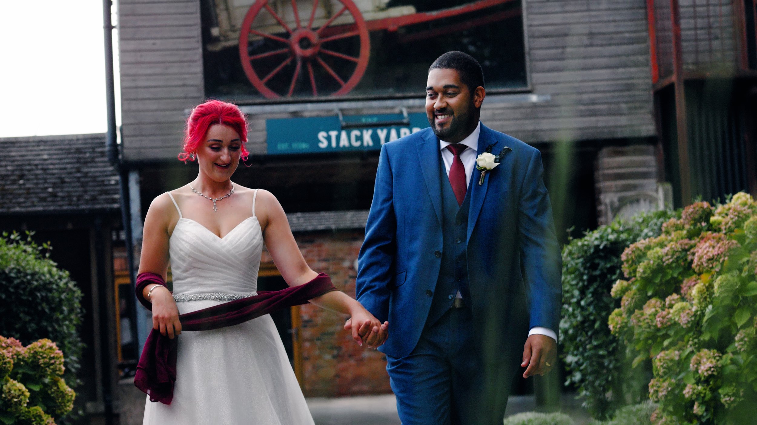 A bride with red hair in a classic white dress with a silver jewelled waistband holds hands with the groom in a blue suit, at the Stack Yard at Donington Park Farmhouse wedding venue. Taken by Derbyshire wedding videographer Laura Baker Productions.